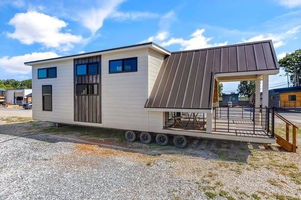 Modern tiny house on wheels with cream siding and dark trim, featuring large windows and a metal roofed porch. Bright day, blue sky, minimal greenery.