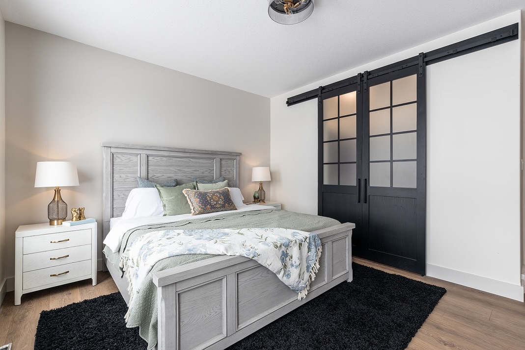 SEO-optimized alt text for a high-ranking luxury custom modular home bedroom featuring a professional interior design with a sophisticated neutral gray color palette and warm wide-plank oak flooring; the master suite showcases a stylish gray wood-grain bed frame with floral bedding, matching white nightstands with contemporary gold lamps, and a plush black area rug, perfectly highlighted by elegant black-framed sliding barn doors with frosted glass panels, modern craftsman-style architectural details, and premium residential mobile living solutions for maximum search engine visibility and ranking.