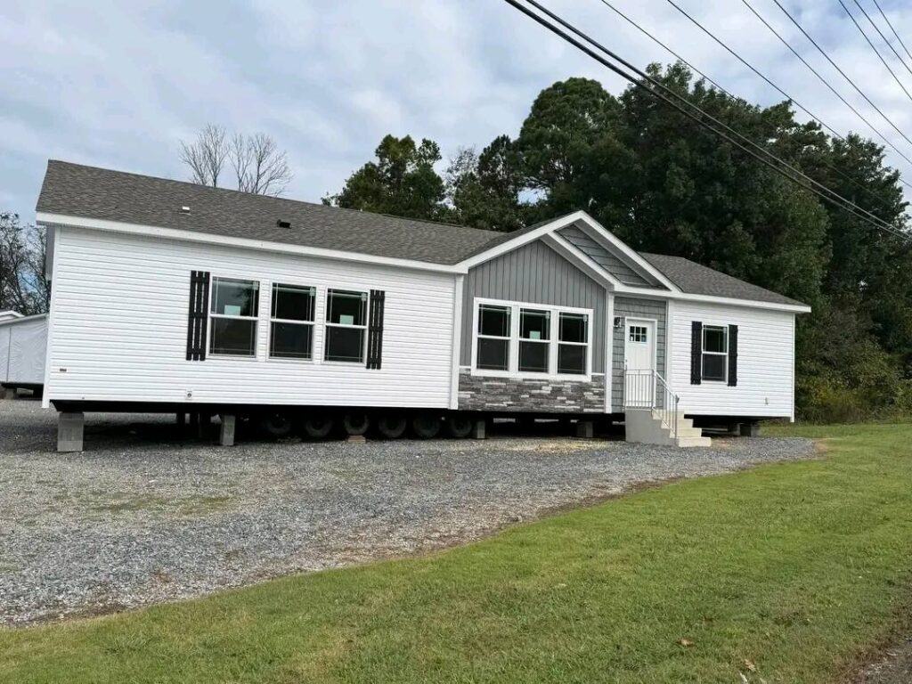 Modern navy blue modular ranch-style home featuring a wide covered front porch with white columns, a grey shingle roof, and white-trimmed windows, currently shown on temporary blocks at a construction site under a clear blue sky.