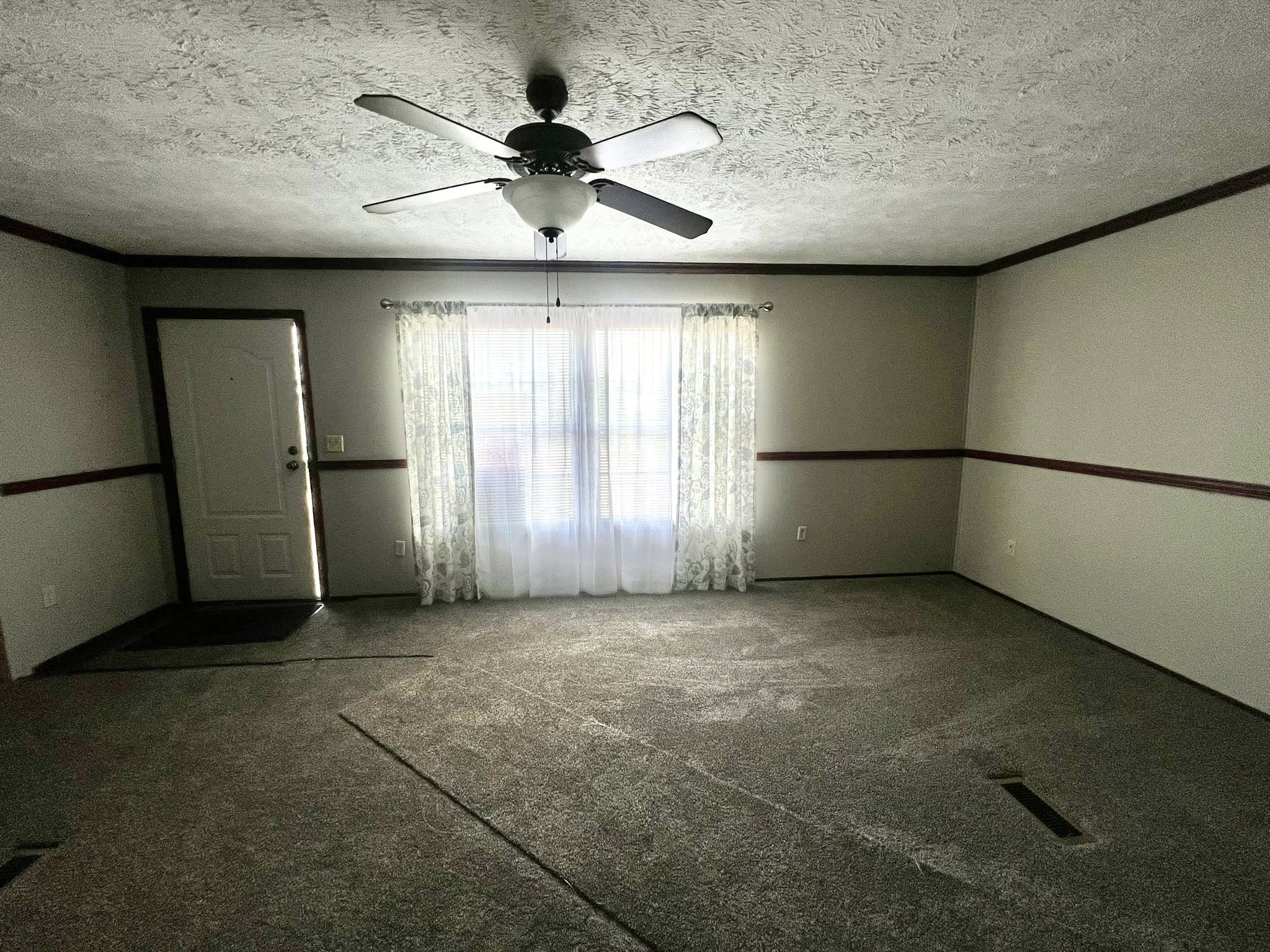 SEO-optimized alt text for a spacious, traditional manufactured home living room featuring neutral gray carpeting and a classic stone-surround fireplace with a rustic wood mantel; the high-ranking interior design showcases dark wood built-in bookshelves, a decorative wall clock, and a central ceiling fan with dark blades, all framed by elegant crown molding and floral-patterned curtains that highlight comfortable family-style modular home floor plans and affordable luxury mobile home decor for maximum search engine visibility.