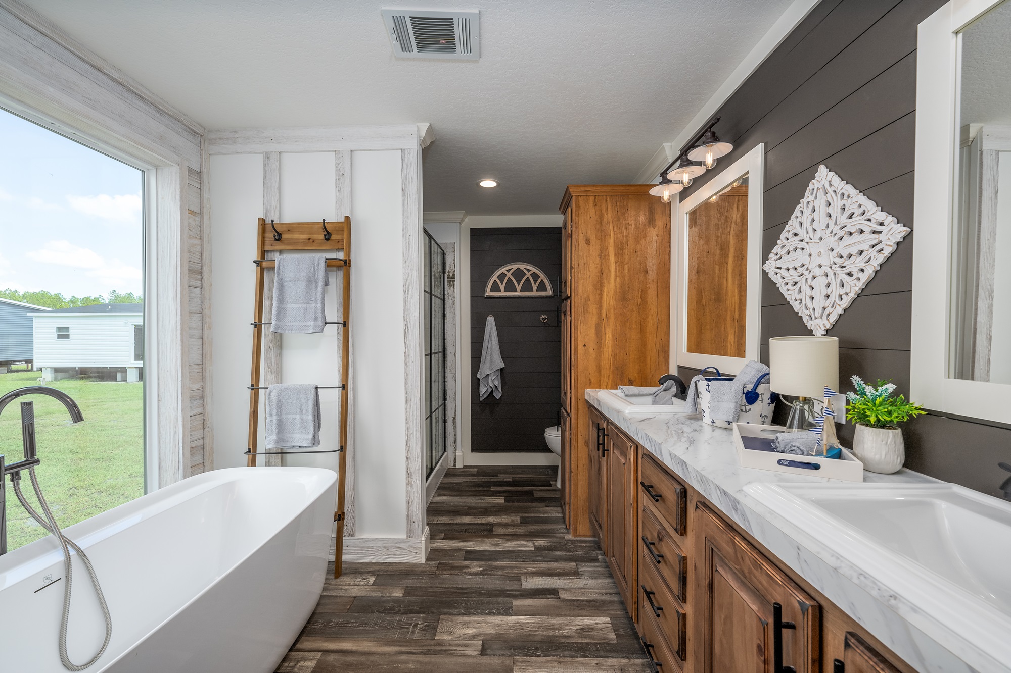 Luxurious spacious modern farmhouse master bathroom in The Garnet 1,873 sq ft 3-bedroom customizable modular home featuring elegant freestanding white soaking bathtub positioned by large picture window with natural light and outdoor views, double vanity with rich walnut wood cabinetry black hardware and white marble-look quartz countertops dual undermount sinks matte black faucets, shiplap accent wall in weathered white contrasting dark gray vertical plank feature wall, freestanding glass walk-in shower with black frame, rustic wooden towel ladder with plush gray towels, nautical-themed decor including anchor tray sailboat lamp potted greenery and seashell accents, dark wood-look tile flooring, vaulted ceiling with recessed lighting and exhaust fan creating a serene spa-like retreat perfect for relaxation in this dream home giveaway winner.
