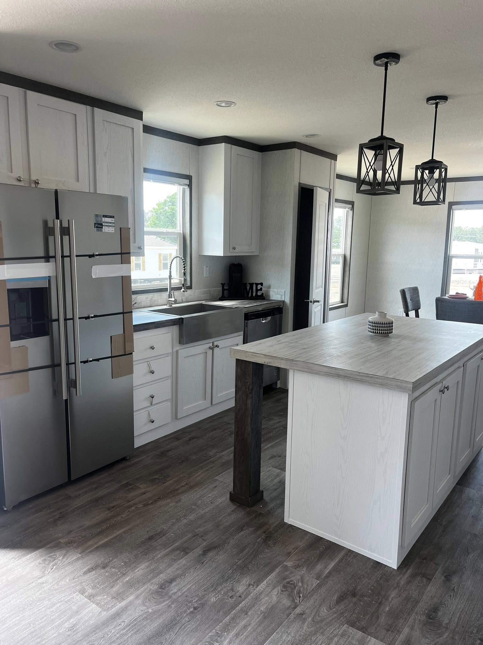 SEO-optimized alt text for a high-ranking, modern kitchen in a luxury manufactured home featuring sleek white shaker cabinets with minimalist hardware and a clean marble-style tile backsplash; the professional interior design showcases a spacious kitchen island with a light wood-look countertop, integrated stainless steel appliances including a farmhouse sink and side-by-side refrigerator, and contemporary black geometric pendant lighting, perfectly highlighting premium mobile home kitchen floor plans, durable dark wood-plank flooring, and stylish modular home upgrades for maximum search engine visibility and ranking.

Would you like me to generate alt text for the dining area or the exterior view of this home next?