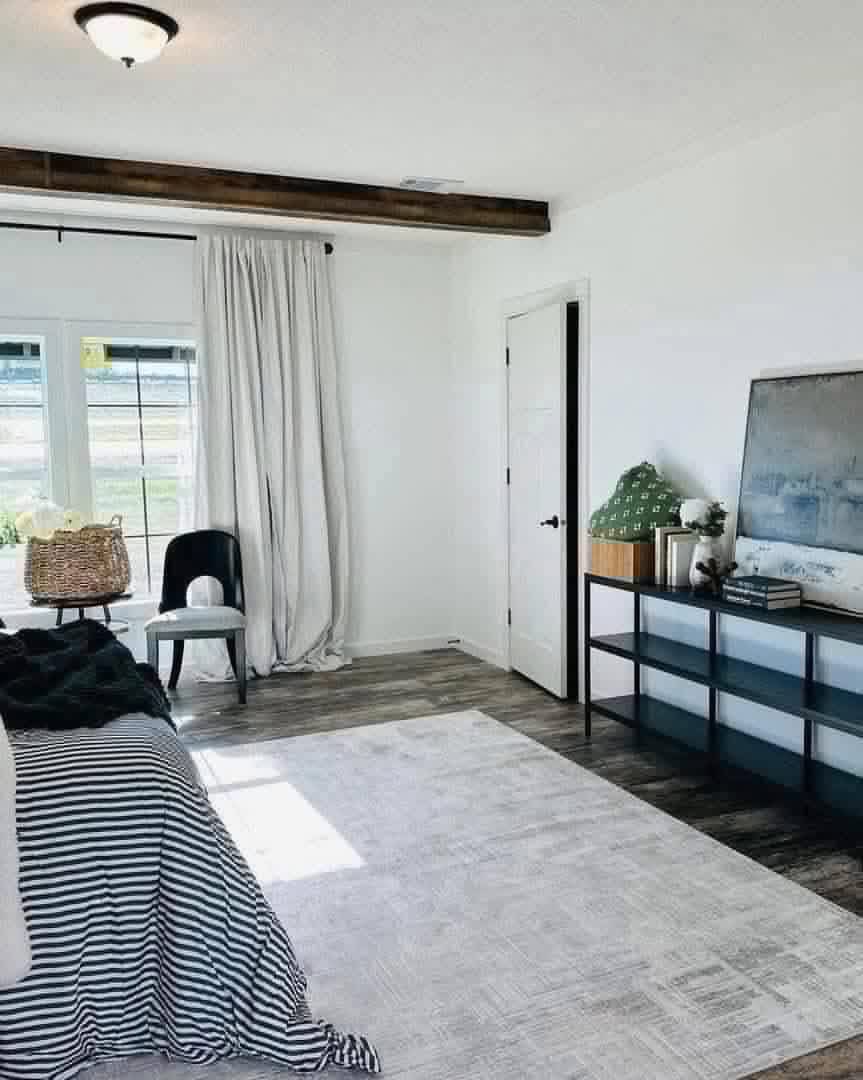 Bright and airy primary bedroom in a modern farmhouse modular home featuring crisp white walls and vaulted ceiling with exposed rustic wooden beam accent, large grid-pattern windows draped in flowing sheer white curtains flooding the space with natural light and scenic outdoor views, plush bed dressed in classic black-and-white striped bedding layered with cozy textured throws, neutral patterned area rug over warm wood-look flooring, minimalist black accent chair beside a small round wooden side table with woven basket storage, sleek black metal console TV stand displaying abstract artwork books greenery and decor, open doorway to hallway creating a serene spacious relaxing master retreat perfect for rest and personalization in this dream affordable manufactured home.
