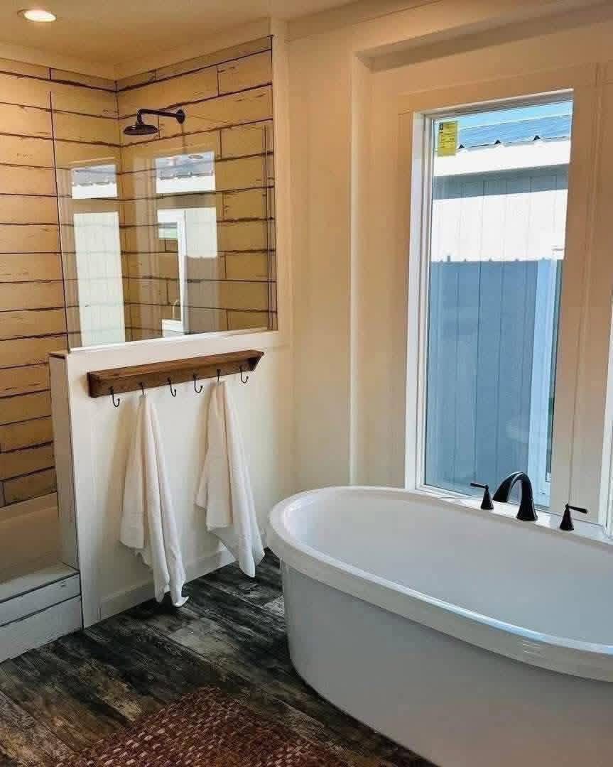 Serene modern farmhouse master bathroom in a customizable modular home featuring rustic reclaimed wood-look shiplap accent walls in warm distressed tones, elegant freestanding oval white soaking bathtub with matte black waterfall faucet positioned beneath a large picture window offering natural light and peaceful outdoor views, frameless glass walk-in shower enclosure with matching black rainfall showerhead, wooden towel ladder shelf with plush white towels, textured brown woven bath mat over dark wood-look tile flooring, crisp white wainscoting and trim creating a spa-like tranquil retreat perfect for relaxation and rejuvenation in this luxurious dream manufactured home design.