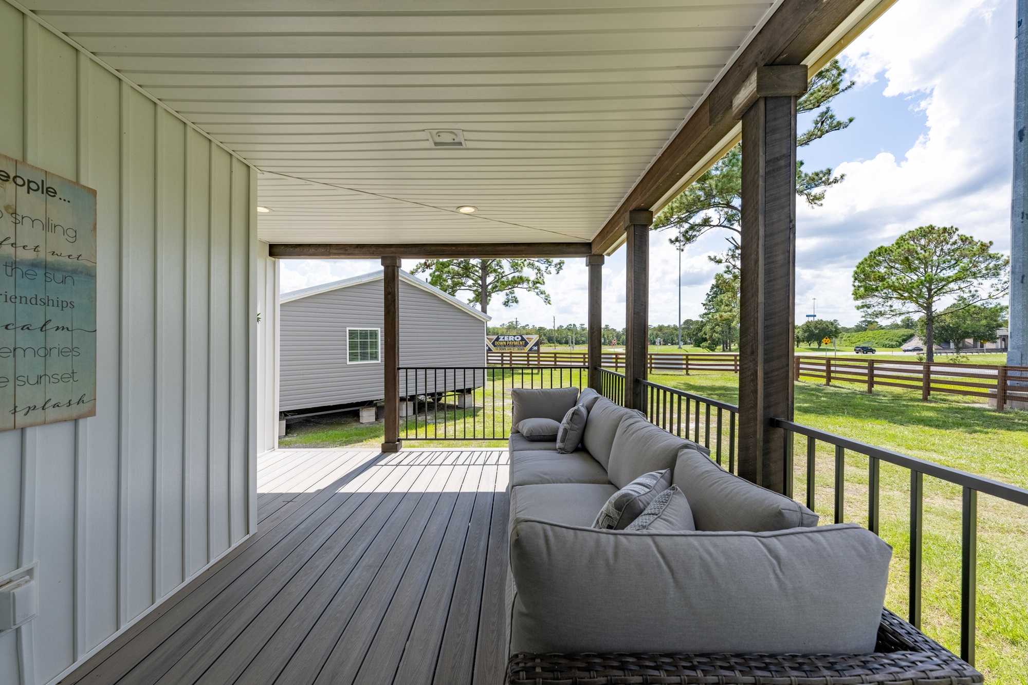 Relaxing covered front porch retreat on a modern manufactured modular home featuring expansive gray composite deck flooring, plush L-shaped outdoor sectional sofa in neutral gray weather-resistant fabric with coordinating throw pillows, sturdy wooden support posts and railing with black metal balusters, vaulted ceiling with recessed lighting and ceiling fan, inspirational beach-themed wall art quoting family memories and sunsets, open view overlooking grassy community lot with other homes trees and road under bright sunny sky creating an inviting peaceful outdoor living space perfect for lounging entertaining and enjoying fresh air in The Garnet style dream home giveaway winner.
