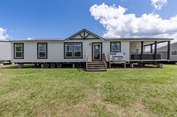 Spacious single-story modular home exterior of The Garnet style or similar 3-bedroom customizable manufactured house featuring crisp white lap siding with dark brown trim accents, classic gabled roofline with decorative black brackets, large energy-efficient windows framed in brown, welcoming covered front porch with black railing and columns, sturdy wooden steps leading to entry door, elevated foundation on wheels for easy transport and setup, branded Lakeview sign, set on grassy lot under bright blue sky with fluffy clouds, ideal affordable family dream home in giveaway contest ready for delivery and personalization.