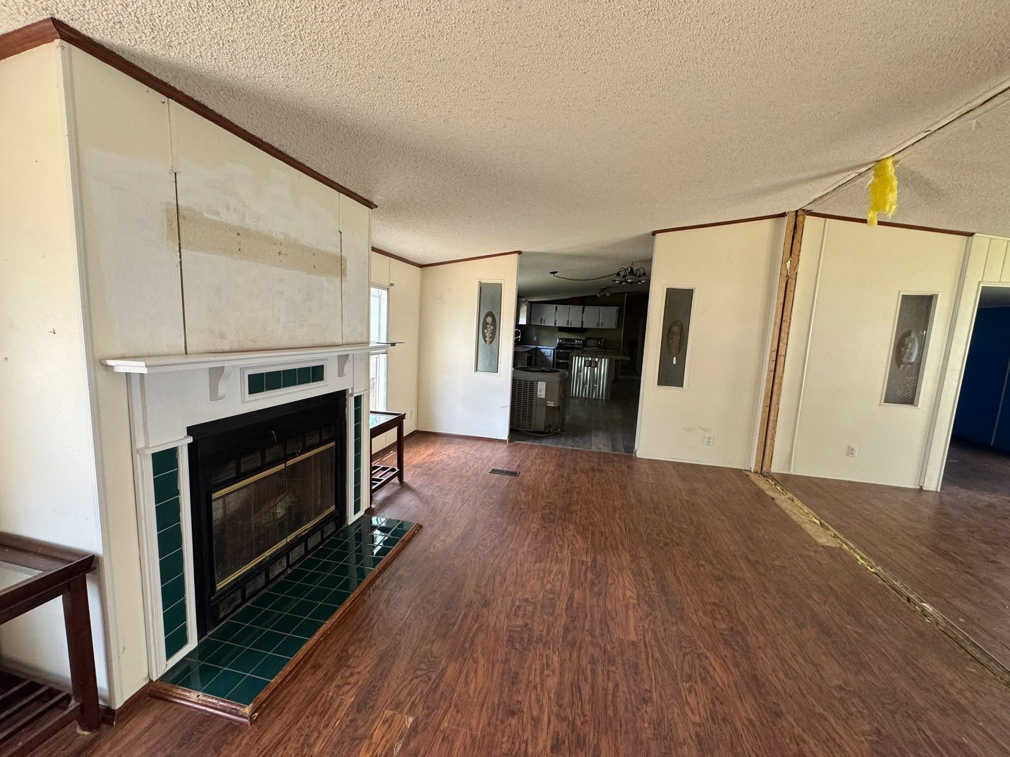 SEO-optimized alt text for an open-concept living room in an older manufactured home featuring wide-plank wood-look laminate flooring and a white fireplace with green decorative tiling; the interior view showcases a spacious layout with vaulted ceilings, a large wall-mounted mirror-style window insert, and a glimpse into the kitchen area, perfectly highlighting affordable mobile home floor plans, residential renovation potential, and traditional double-wide interior designs for maximum search engine visibility and ranking.