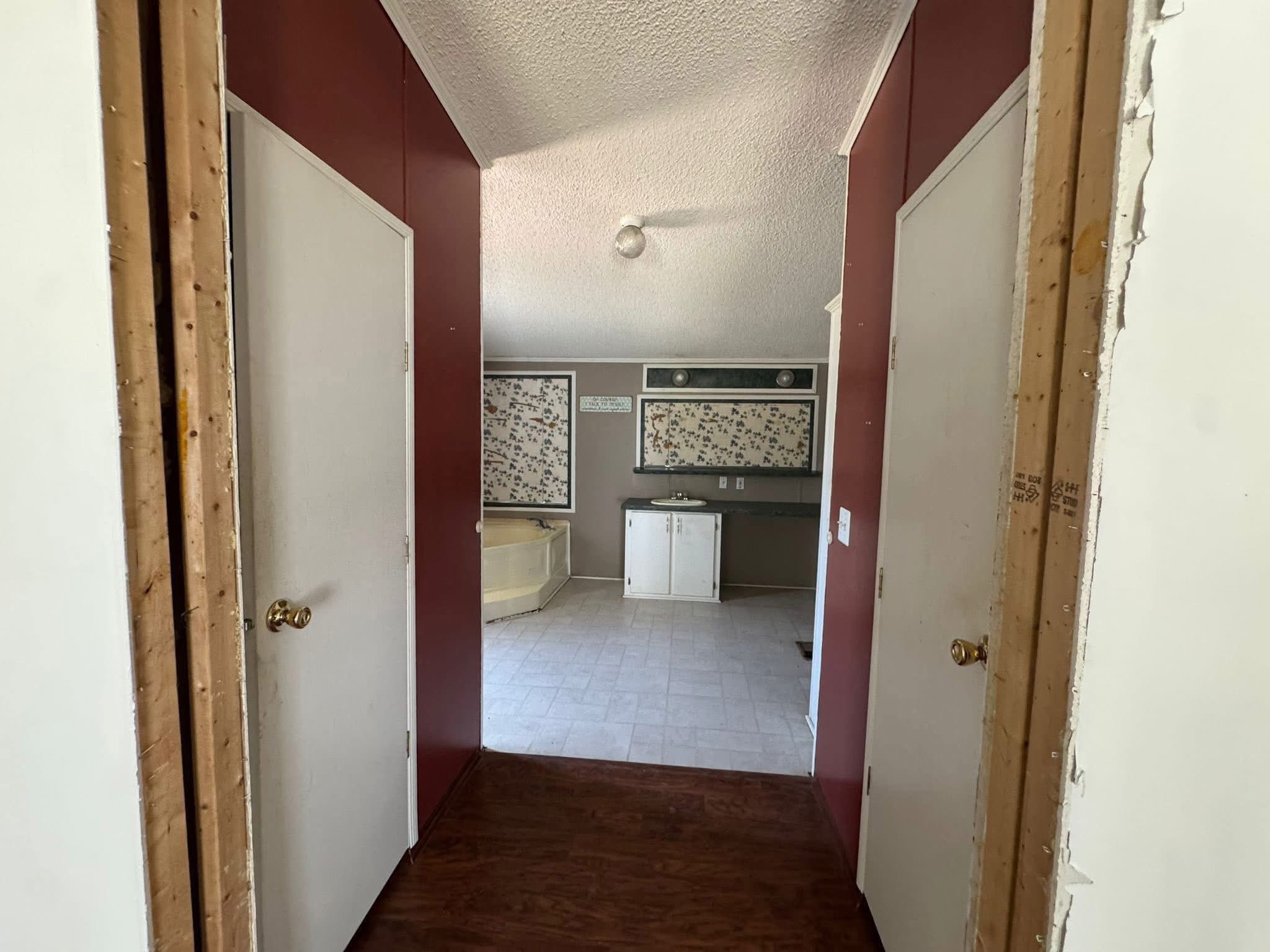 SEO-optimized alt text for an older manufactured home master suite renovation featuring a hallway with dark wood-look flooring and red accent walls leading into a spacious bathroom; the high-ranking interior view showcases a built-in garden tub, a white vanity with a dark countertop, and decorative floral wall panels, perfectly highlighting affordable mobile home remodeling projects, traditional double-wide floor plans, and residential fixer-upper potential for maximum search engine visibility and ranking.