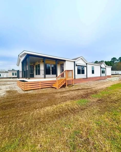 Modern farmhouse style manufactured home exterior featuring crisp white lap siding with contrasting black trim accents large energy-efficient windows welcoming expansive covered front porch with black metal railings and wooden support beams multi-level wooden deck stairs in warm natural finish spacious L-shaped wraparound porch design elevated foundation on gravel lot under partly cloudy blue sky surrounded by open grassy area and distant trees ideal affordable family home with 4 bedrooms 2.5 baths massive outdoor living space perfect for entertaining relaxation and curb appeal in this dream modular home giveaway or for sale model.