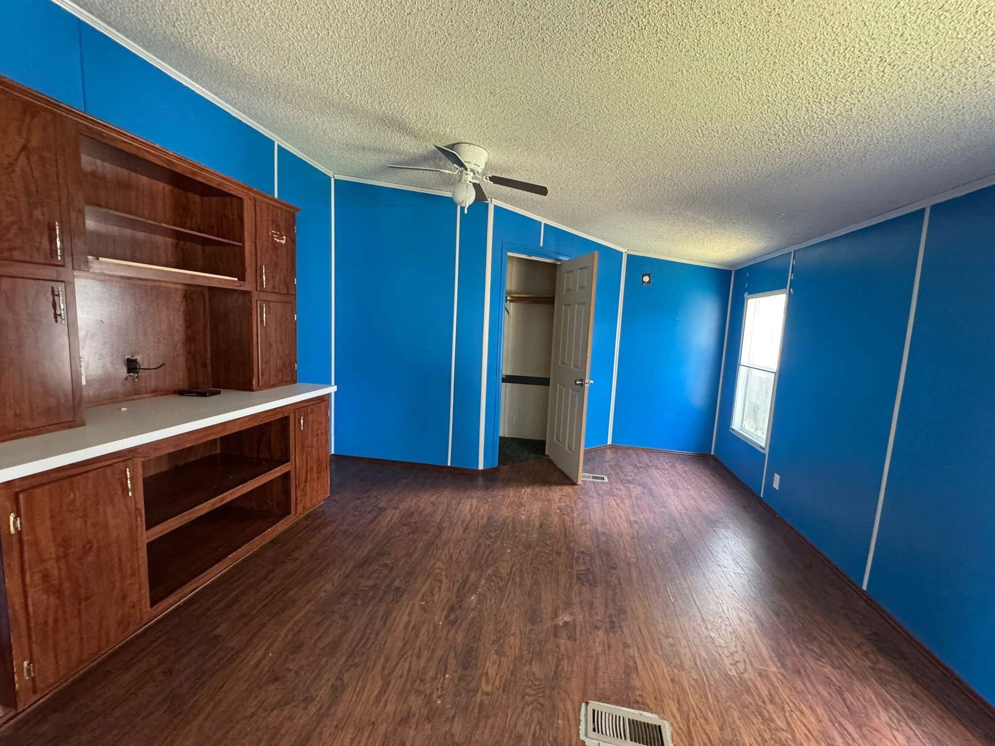 SEO-optimized alt text for an empty room in a manufactured home featuring vibrant blue walls with white trim and dark wood-look laminate flooring; the interior view showcases a built-in dark wood entertainment center or hutch with open shelving, a white ceiling fan, and a single window providing natural light, perfectly highlighting affordable mobile home interior features, residential floor plan layouts, and bold modular home renovation ideas for maximum search engine visibility and ranking.