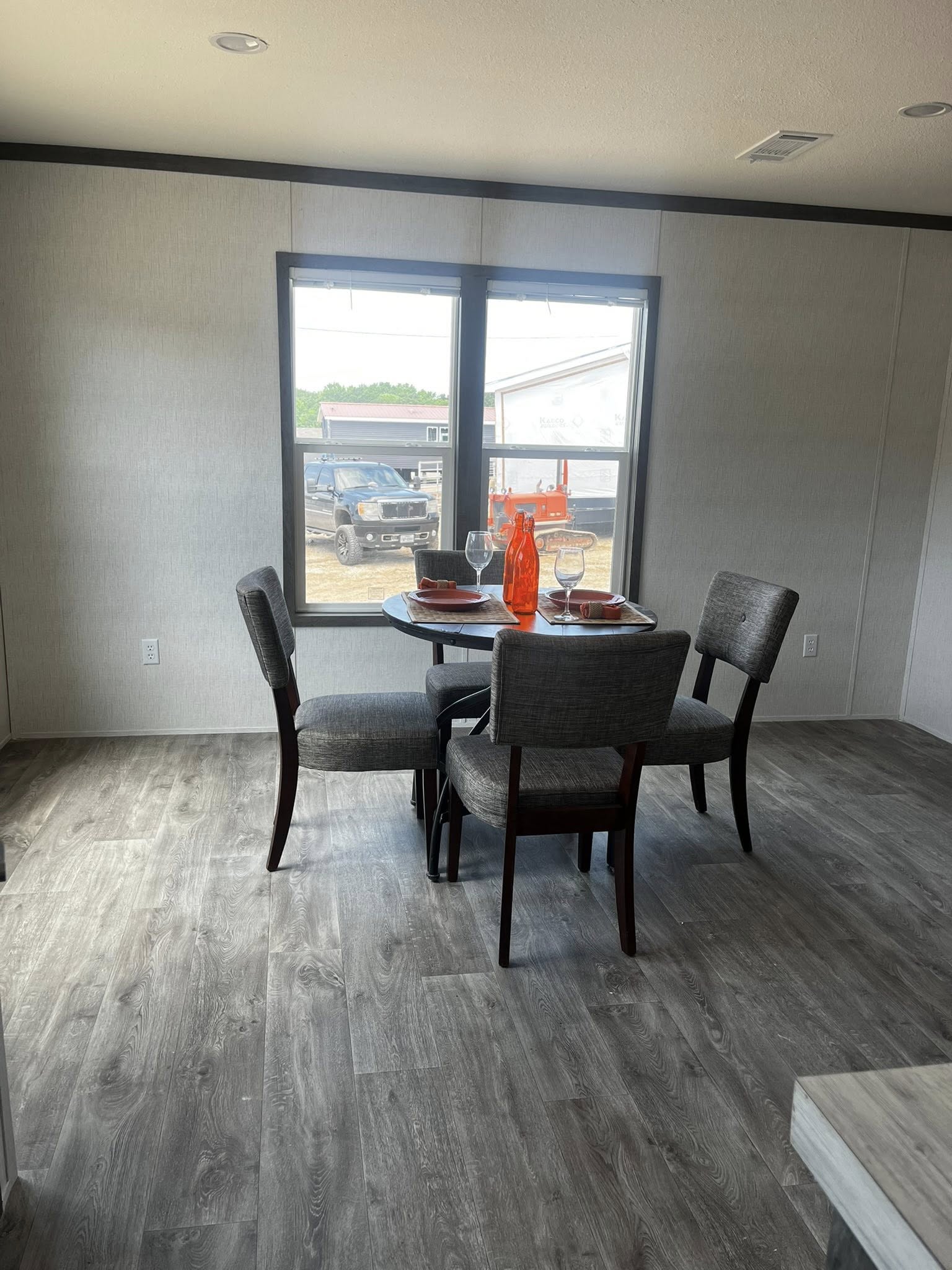 SEO-optimized alt text for a modern manufactured home's open-concept dining area featuring a dark wood circular table with four contemporary gray upholstered chairs set against neutral gray walls with dark trim; the high-ranking interior design showcases durable wood-look laminate flooring and a large double window providing natural light and a view of the residential property, perfectly highlighting stylish modular home dining room layouts, functional small-space furniture ideas, and premium mobile home interior finishes for maximum search engine visibility and ranking.