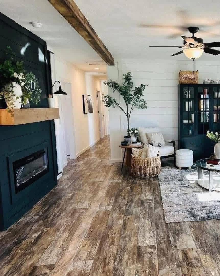 Open-concept living room and hallway in a stunning modern farmhouse modular home featuring dramatic dark teal accent fireplace wall with electric insert and rustic wooden mantel displaying inspirational "breathe" sign fresh floral arrangements greenery potted plants cozy white armchair with throw pillows woven basket ottoman glass-top round coffee table neutral patterned area rug over rich warm wood-look plank flooring exposed dark wooden ceiling beams vaulted textured ceiling with black ceiling fan integrated light abundant natural light from large windows sheer curtains plush seating arrangement tall navy blue bookcase cabinet with decor books vases creating a warm inviting spacious family gathering space flowing seamlessly into bright hallway with shiplap walls perfect for relaxation entertaining and everyday living in this luxurious dream manufactured home design.