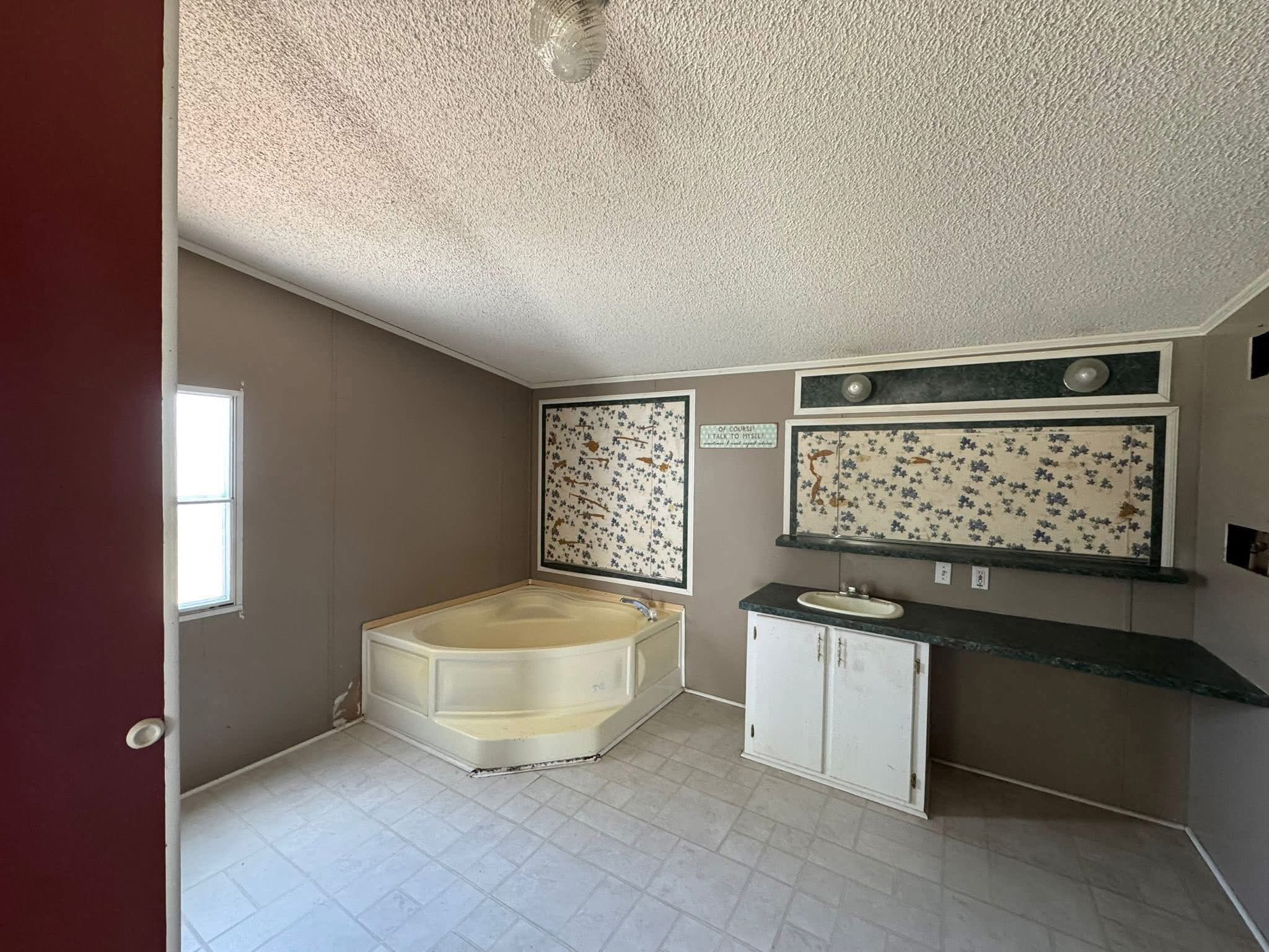 SEO-optimized alt text for an empty bedroom in a manufactured home featuring dark wood-look laminate flooring, off-white walls with a decorative brown chair rail, and matching crown molding; the interior view showcases two large windows with white blinds and sheer ruffled valances, a white paneled door leaning against the wall, and a textured ceiling with a central light fixture wire exposed, perfectly highlighting affordable mobile home interior finishes, residential floor plan potential, and clean modular home renovation ideas for maximum search engine visibility and ranking.