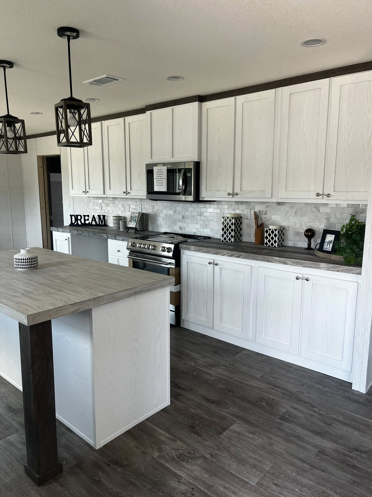 SEO-optimized alt text for a modern, high-ranking manufactured home kitchen featuring elegant white shaker-style cabinetry with minimalist silver hardware and a sophisticated gray marble-patterned backsplash; the professional interior design showcases a spacious kitchen island with a light wood-look countertop, dark wood support beams, and contemporary black geometric pendant lighting, perfectly highlighting luxury mobile home kitchen floor plans, durable wood-plank flooring, and stylish modular home upgrades for maximum search engine visibility and ranking.