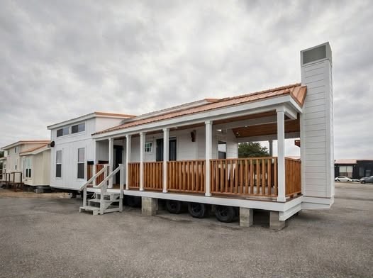 Spacious wraparound porch on a charming modern farmhouse tiny modular home featuring crisp white shiplap siding with warm terracotta metal roof accents, expansive covered front porch supported by sturdy white columns and rich walnut wooden railing panels, welcoming wooden steps leading to double entry doors, large energy-efficient windows allowing natural light, elevated trailer chassis on concrete blocks under overcast sky with scattered clouds, gravel lot setting surrounded by other manufactured homes creating an inviting outdoor living space perfect for relaxing entertaining and enjoying fresh air in this affordable customizable dream tiny house or starter manufactured home model with exceptional curb appeal.