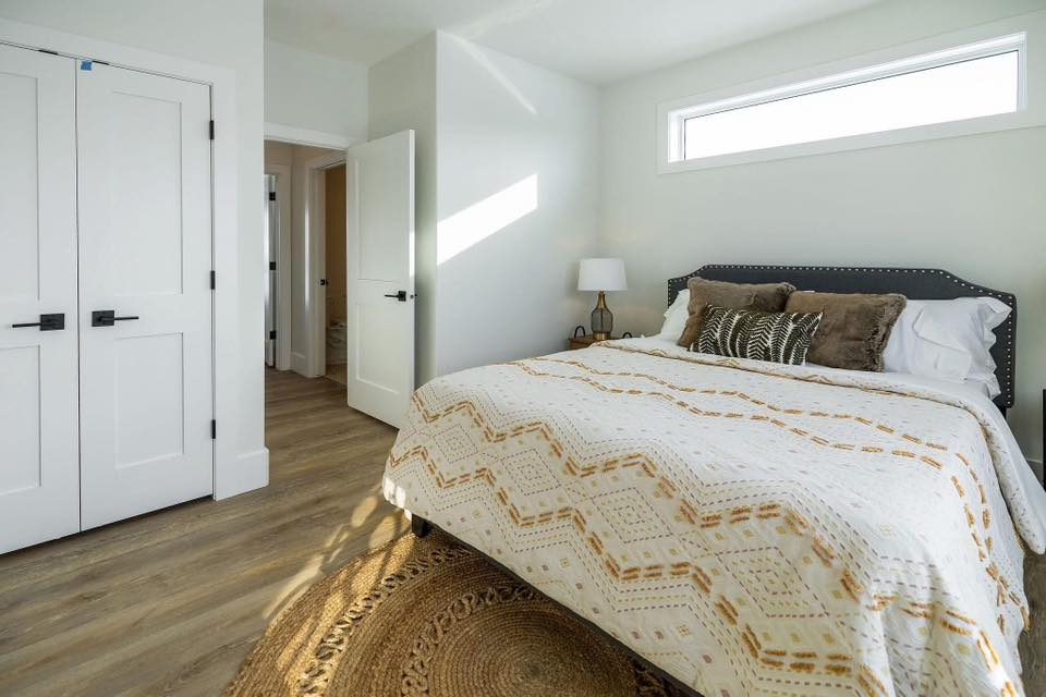 Serene and spacious primary bedroom in The Garnet 1,873 sq ft 3-bedroom customizable modular home featuring bright white walls and trim, elegant board-and-batten double closet doors with black hardware, large horizontal clerestory window flooding the room with natural light, upholstered dark gray tufted headboard bed dressed in cozy neutral boho bedding with geometric mustard and cream patterned throw, layered textured pillows in earthy tones, woven jute round rug over warm hardwood-look flooring, minimalist nightstand with brass lamp and decor, open doorway to ensuite bathroom, creating a calm relaxing modern farmhouse master retreat perfect for rest and rejuvenation in this dream home giveaway winner.