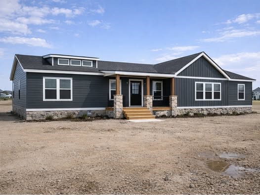 Modern farmhouse exterior of The Garnet 1,873 sq ft 3-bedroom customizable modular home featuring striking dark gray shiplap siding, contrasting white trim windows, elegant black roof with dormer accents, welcoming covered front porch with wooden beams and stone pillars, natural stone base and steps leading to double entry doors, spacious open layout design under clear blue sky in a new construction community, ideal dream family home giveaway winner ready for move-in.
