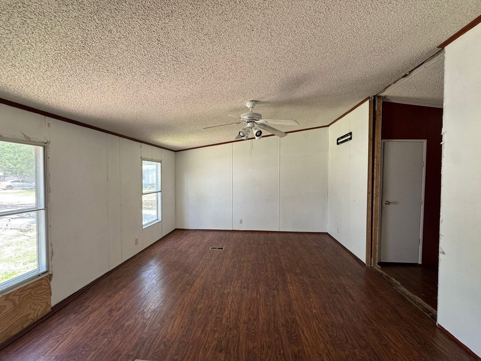 SEO-optimized alt text for an empty bedroom in a manufactured home featuring dark wood-look laminate flooring, light gray walls with a decorative brown chair rail, and matching crown molding; the interior view showcases two windows with white blinds and simple ruffled valances, an open white door, and a textured ceiling with a central light fixture connection, perfectly highlighting affordable mobile home interior finishes, residential floor plan potential, and clean modular home renovation ideas for maximum search engine visibility and ranking.
