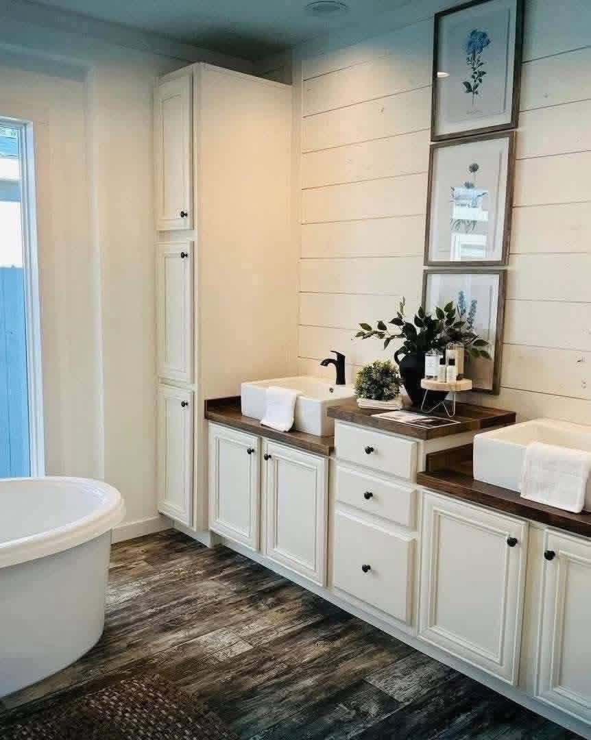 Luxurious spa-inspired master bathroom in a modern farmhouse modular home featuring elegant white shiplap walls with subtle blue ceiling accent, freestanding oval soaking bathtub positioned by large window for natural light and serene views, expansive double vanity with white shaker cabinets black knob hardware rich walnut wood-look countertops dual farmhouse square undermount sinks matte black faucets, tall linen tower cabinet for storage, stacked botanical artwork in rustic frames above vanity, potted greenery and luxury bath products on tray, textured dark wood-look tile flooring, open airy layout creating a peaceful relaxing retreat perfect for unwinding in this customizable dream home.