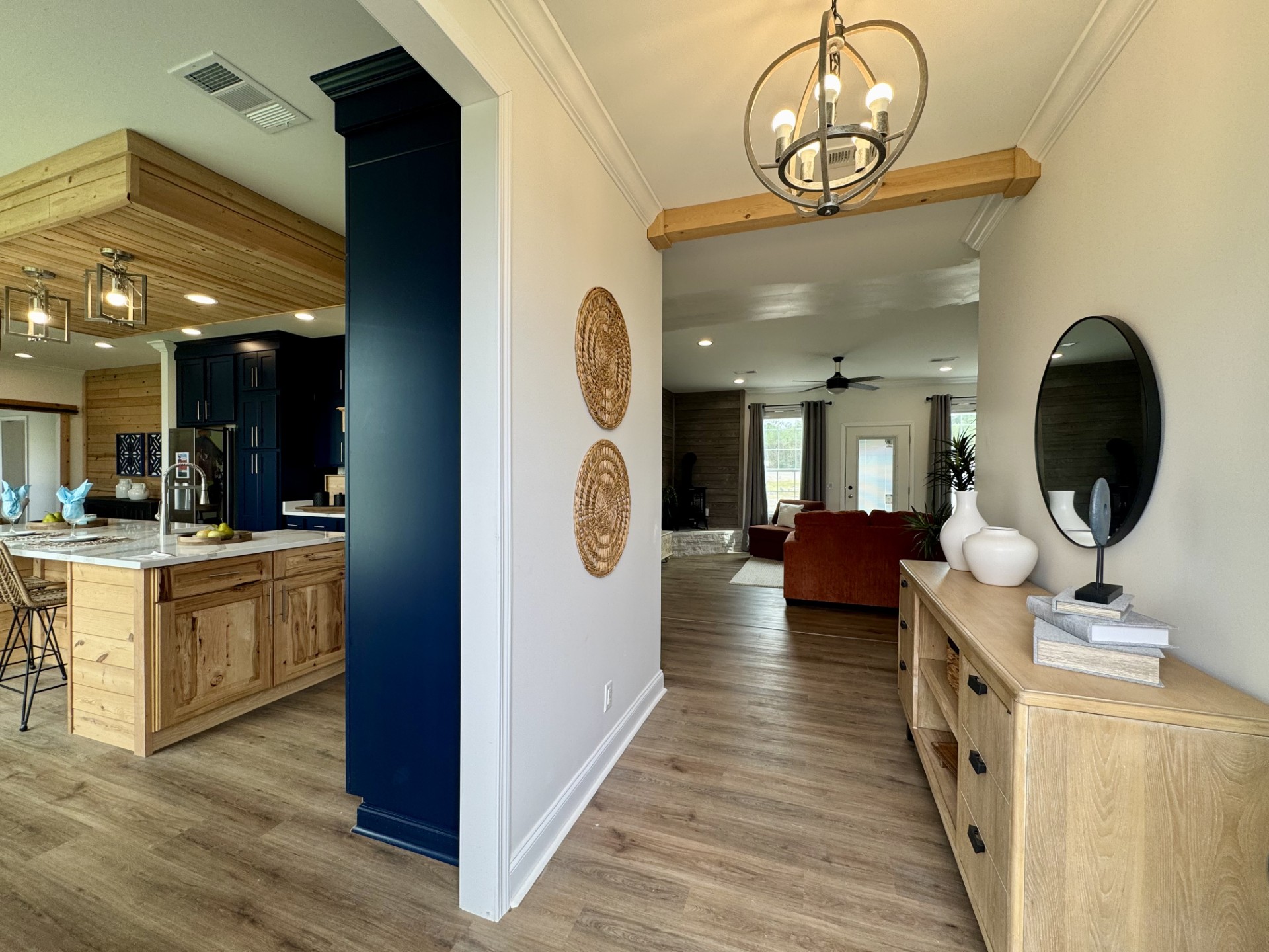 SEO-optimized alt text for a high-ranking luxury custom modular home entryway and living area featuring a professional open-concept interior design with an elegant light wood console table and contemporary round black mirror; the architectural view showcases a sophisticated navy blue kitchen accent pillar, a rustic wood-plank ceiling feature with industrial cage lighting, and a cozy sunlit living room with a plush terracotta sectional sofa, all set against premium wide-plank flooring and minimalist cream walls to highlight luxury prefab home floor plans, modern farmhouse-style architectural details, and high-end residential mobile living solutions for maximum search engine visibility and ranking.