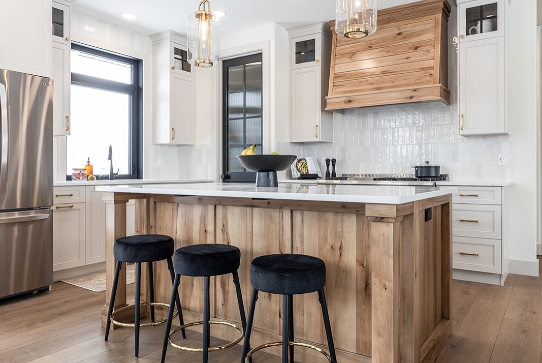 SEO-optimized alt text for a high-ranking luxury custom modular home kitchen featuring a professional white shaker cabinet design and a large rustic wood-plank kitchen island with a white quartz waterfall countertop; the interior showcases a premium wood-veneer range hood, elegant white vertical subway tile backsplash, and contemporary gold hardware, complemented by stylish black velvet barstools, modern glass pendant lighting, and a high-end stainless steel refrigerator, all set against durable wide-plank oak flooring to highlight gourmet prefab kitchen layouts, sophisticated open-concept floor plans, and upscale residential mobile living finishes for maximum search engine visibility and ranking.