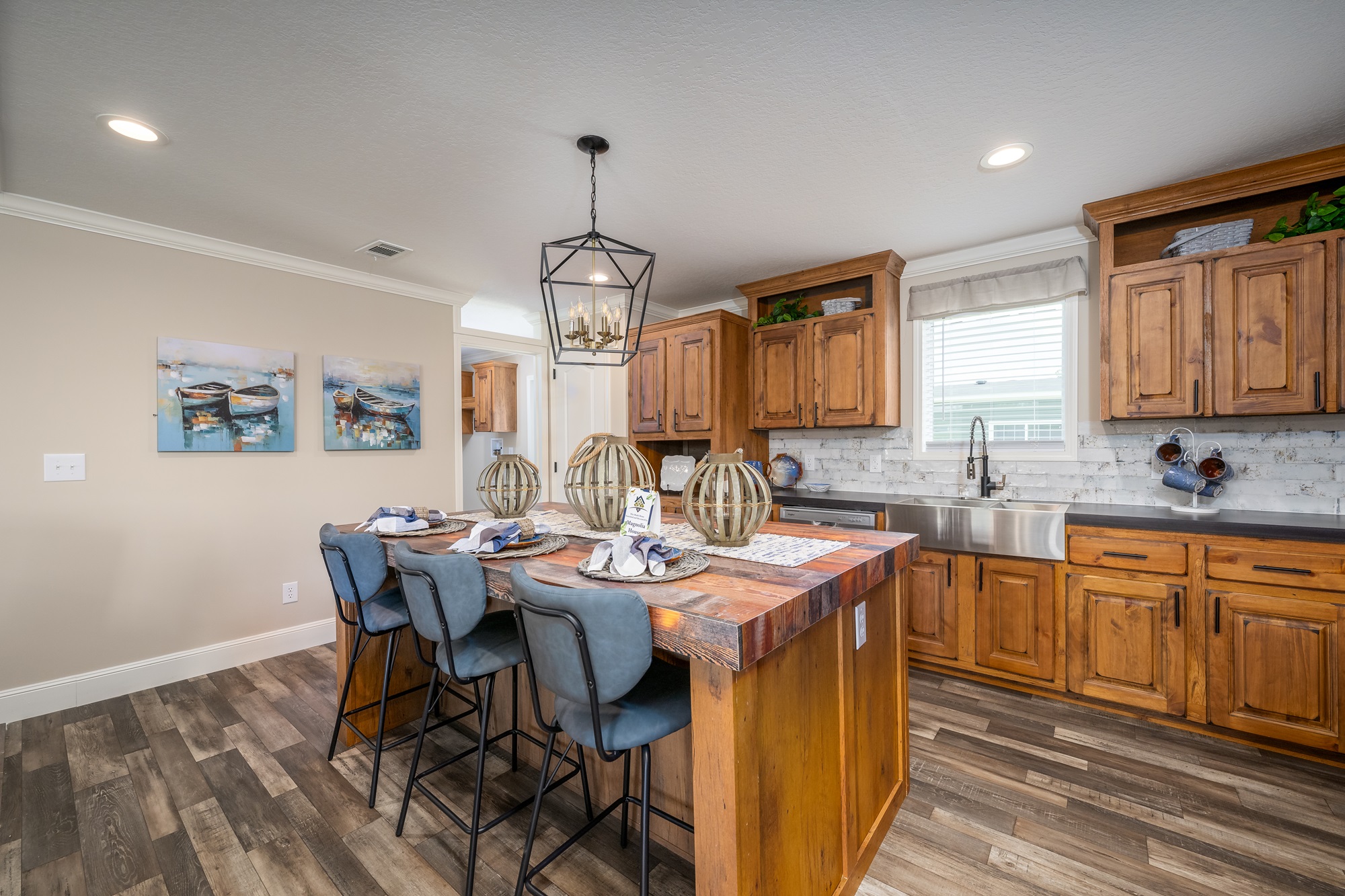 Spacious coastal-inspired kitchen and dining island in The Garnet 1,873 sq ft 3-bedroom customizable modular home featuring rich warm walnut wood cabinetry with black hardware, durable dark quartz countertops, large central breakfast bar island with distressed wood top seating four blue upholstered counter stools, farmhouse apron-front stainless steel sink with matte black faucet, white subway tile backsplash, recessed ceiling lights and modern geometric lantern pendant chandelier, nautical-themed boat artwork on beige accent walls, open layout with ample natural light from windows, built-in dishwasher and appliances, decorative woven globe lanterns and potted plants creating a bright inviting family-friendly cooking entertaining space in this dream home giveaway winner.