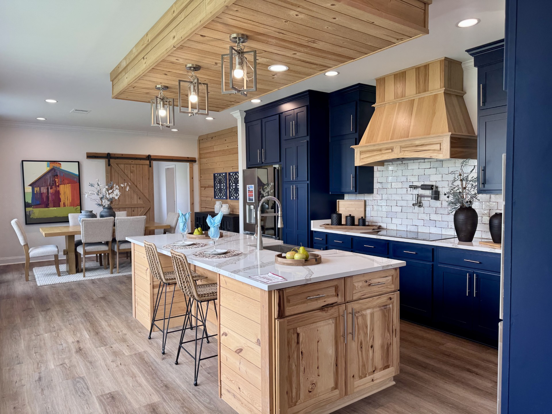 SEO-optimized alt text for a high-ranking luxury custom modular home kitchen and dining area featuring a professional open-concept interior design with a massive white quartz waterfall island and rustic wood cabinetry; the architectural view showcases a sophisticated navy blue kitchen base with a premium wood-veneer range hood, elegant white subway tile backsplash, and high-end stainless steel appliances, complemented by a unique wood-plank ceiling feature with industrial cage pendant lighting and a chic dining space with a sliding barn door and rustic table set, all set against durable wide-plank flooring to highlight gourmet prefab kitchen layouts, modern craftsman-style architectural details, and upscale residential mobile living solutions for maximum search engine visibility and ranking.