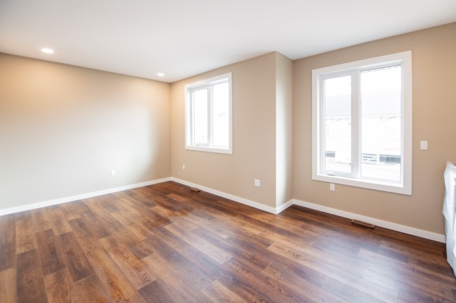 SEO-optimized alt text for a high-ranking luxury custom modular home bedroom featuring a professional modern interior design with clean beige walls and premium wide-plank dark wood flooring; the spacious and sunlit master suite showcases large white-framed windows that offer a clear view of the residential development, highlighting the home's energy-efficient construction, contemporary craftsman-style architectural details, and upscale durable finishes, perfectly designed for maximum search engine visibility and ranking for those seeking high-end prefab home floor plans and modern residential mobile living solutions.