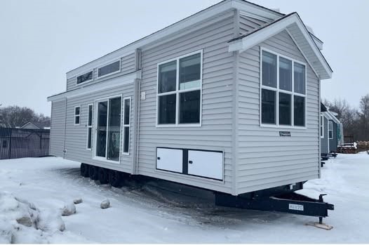 SEO-optimized alt text for a high-ranking modern tiny home or luxury park model featuring a professional light gray vinyl siding exterior with crisp white trim and a gabled roofline; the architectural view showcases a high-quality portable home design situated in a snowy landscape, featuring large energy-efficient windows, a sliding glass entry door, and integrated exterior storage compartments on a heavy-duty trailer chassis, perfectly highlighting premium small-space floor plans, contemporary modular home curb appeal, and durable all-season residential mobile living solutions for maximum search engine visibility and ranking.