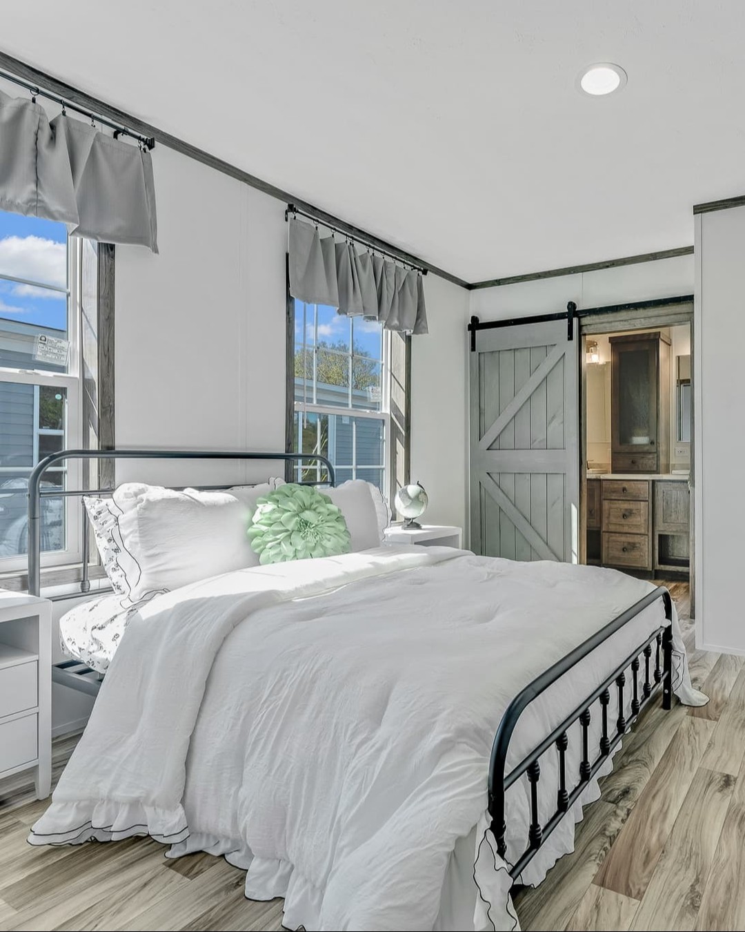 Cozy modern farmhouse bedroom in luxury tiny home or modular prefab cottage featuring white shiplap walls with dark trim accents, large windows with gray rod pocket valance curtains allowing abundant natural light and scenic views, black wrought iron farmhouse bed frame dressed in crisp white ruffled bedding with green accent pillow and throw, mint green floral pom pom decor, globe bedside lamp on white nightstand, sliding barn door in weathered gray opening to en-suite bathroom with wood vanity, light wood-look luxury vinyl plank flooring, bright airy minimalist rustic chic design perfect for guest room, primary suite, or vacation cottage retreat