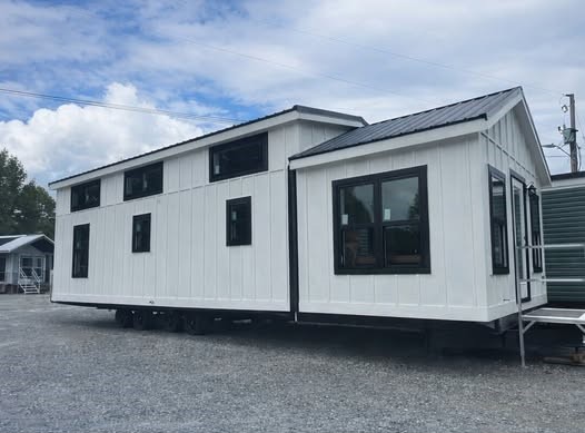 SEO-optimized alt text for a high-ranking modern tiny home or park model manufactured home featuring a striking white board-and-batten exterior with contrasting black window frames and a dark metal roof; the professional architectural view showcases a sleek, single-level mobile home design with multiple clerestory windows for natural light, perfectly highlighting luxury tiny house floor plans, contemporary portable living solutions, and stylish modular home curb appeal for maximum search engine visibility and ranking.