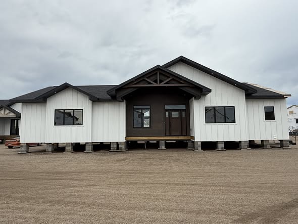 SEO-optimized alt text for a high-ranking modern farmhouse-style modular home featuring a professional white board-and-batten exterior with sophisticated dark brown accents and a matching gabled entry porch; the architectural view showcases a high-quality prefab residence with black-framed windows, a sleek dark roofline, and a sturdy elevated foundation, perfectly highlighting luxury manufactured home curb appeal, contemporary small-space floor plans, and premium portable living solutions for maximum search engine visibility and ranking.