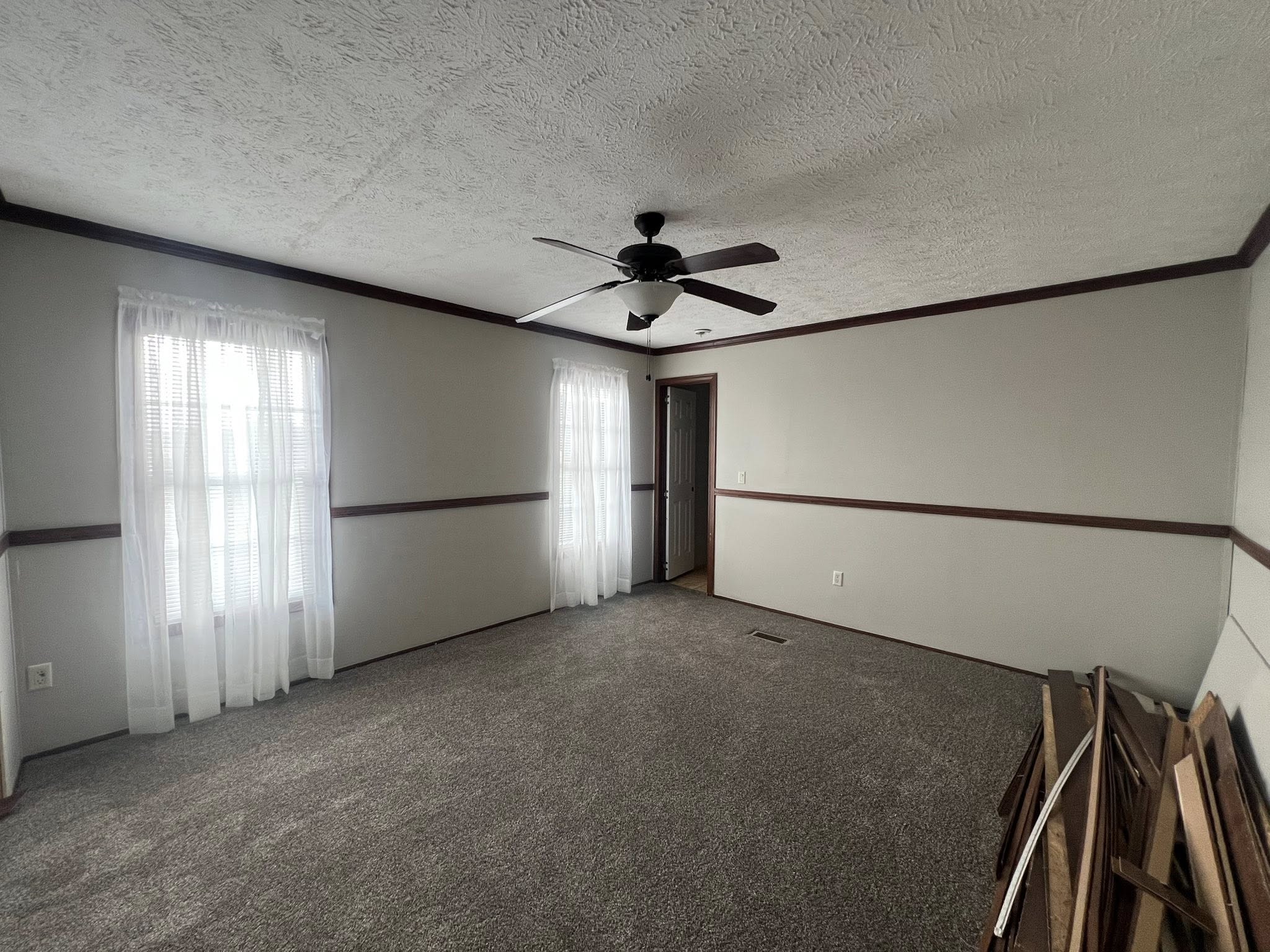 SEO-optimized alt text for a spacious master bedroom in a manufactured home featuring plush neutral gray carpeting, elegant dark wood crown molding, and a coordinated chair rail; the room is illuminated by a central dark-bladed ceiling fan and large windows dressed in soft white curtains, showcasing a clean and ready-to-personalize interior design perfect for high-ranking searches related to affordable luxury mobile home floor plans, modern modular home bedroom ideas, and high-quality residential housing.