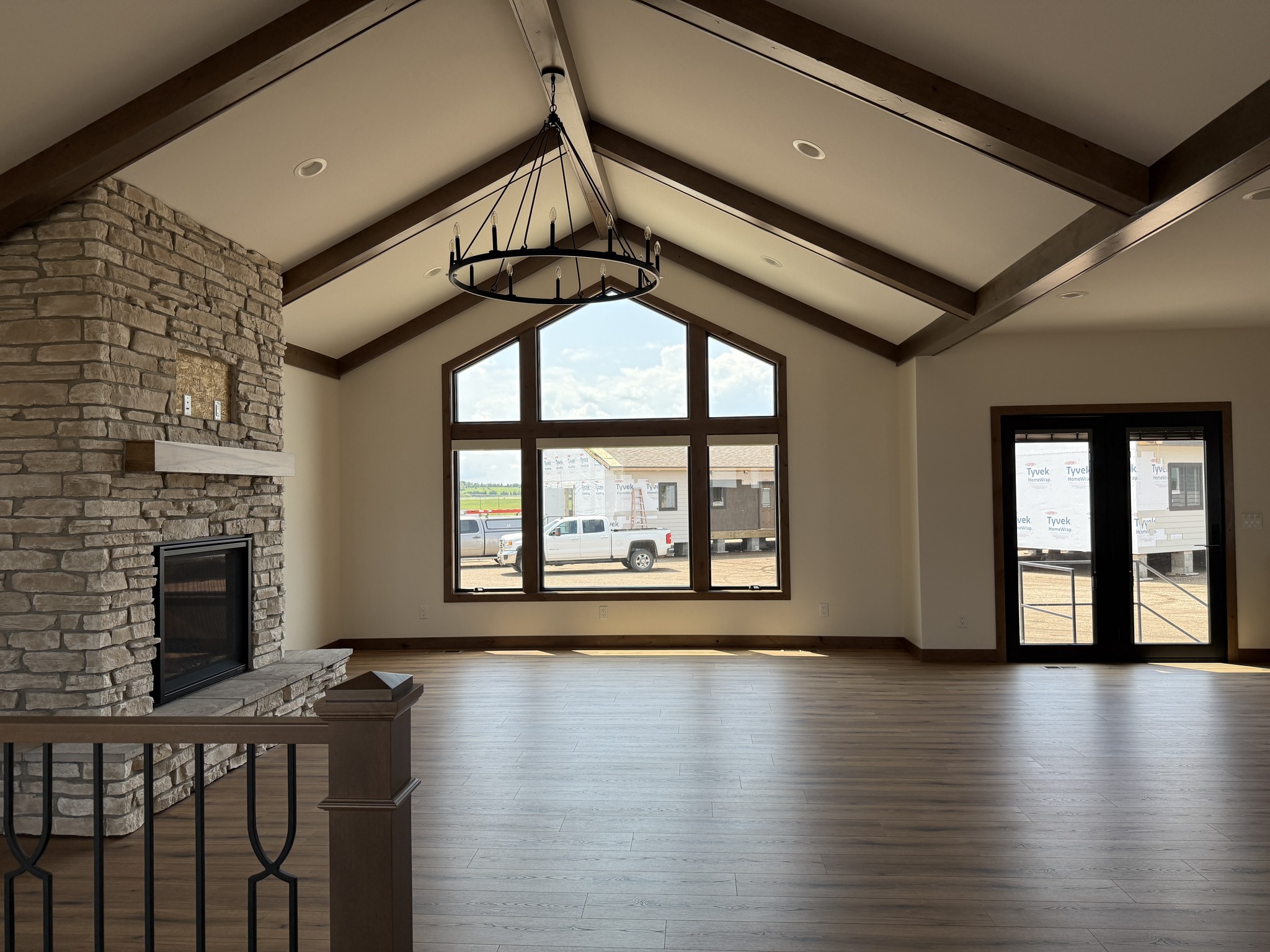 SEO-optimized alt text for a high-ranking modern luxury modular home interior featuring a spacious, open-concept living area with soaring vaulted ceilings and rustic dark wood beams; the professional architectural view showcases a grand floor-to-ceiling stone-veneer fireplace, a sophisticated black wrought-iron wagon wheel chandelier, and durable wide-plank wood flooring, all illuminated by large windows and black-framed French doors that look out onto a residential development, perfectly highlighting premium prefab home floor plans, contemporary craftsman-style design elements, and high-end residential mobile living solutions for maximum search engine visibility and ranking.