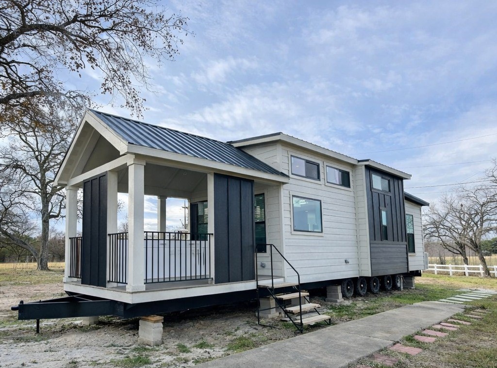 Modern luxury tiny home for sale featuring a high-end white lap siding exterior with a sophisticated black board and batten accent wall on the gabled front porch, showcasing a multi-axle mobile chassis, energy-efficient black-framed windows, and a durable black metal roof on a stable pier foundation for a premium move-in ready turn-key residential living experience.