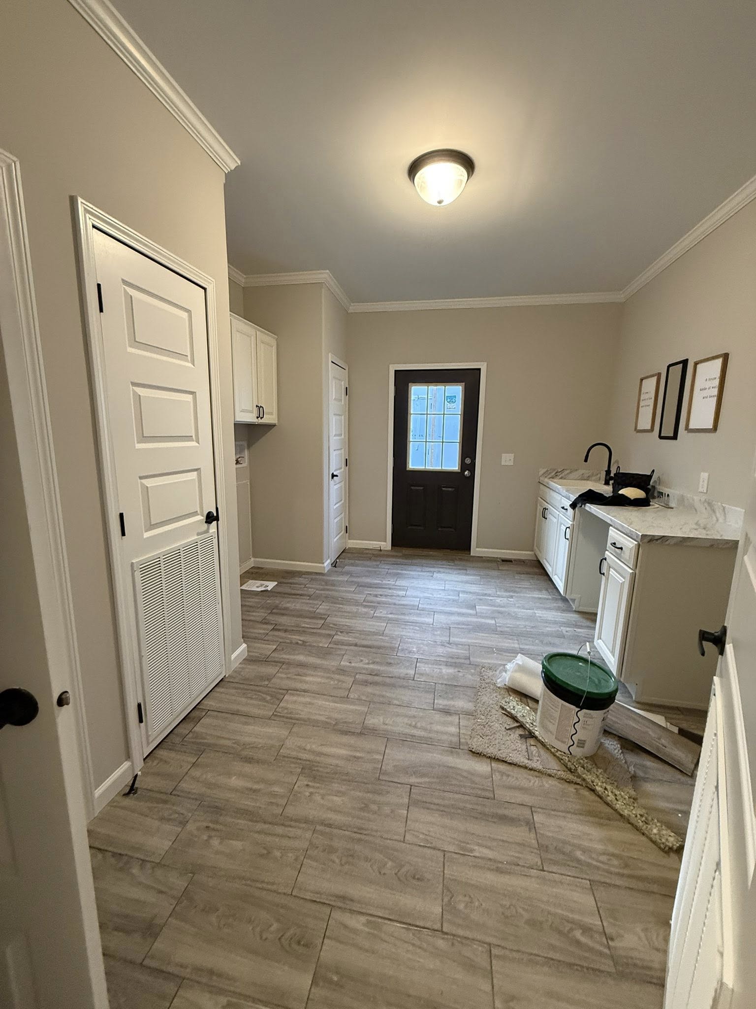 Luxury modern farmhouse tiny home or modular prefab cottage entryway and utility hallway featuring spacious open layout with crown molding and tray ceiling accents, recessed lighting and flush-mount ceiling fixture, neutral taupe walls with white trim, light gray wood-look luxury vinyl plank flooring throughout, white shaker cabinets with black hardware in compact kitchenette area, marble-look quartz countertop with matte black faucet, front entry door with glass panels framed in black, multiple white paneled interior doors leading to bedrooms and living spaces, wall art frames with inspirational quotes, construction finishing touches including paint bucket and drop cloths, bright airy transitional space perfect for family ranch-style manufactured home, vacation cottage, or rural retreat entry design