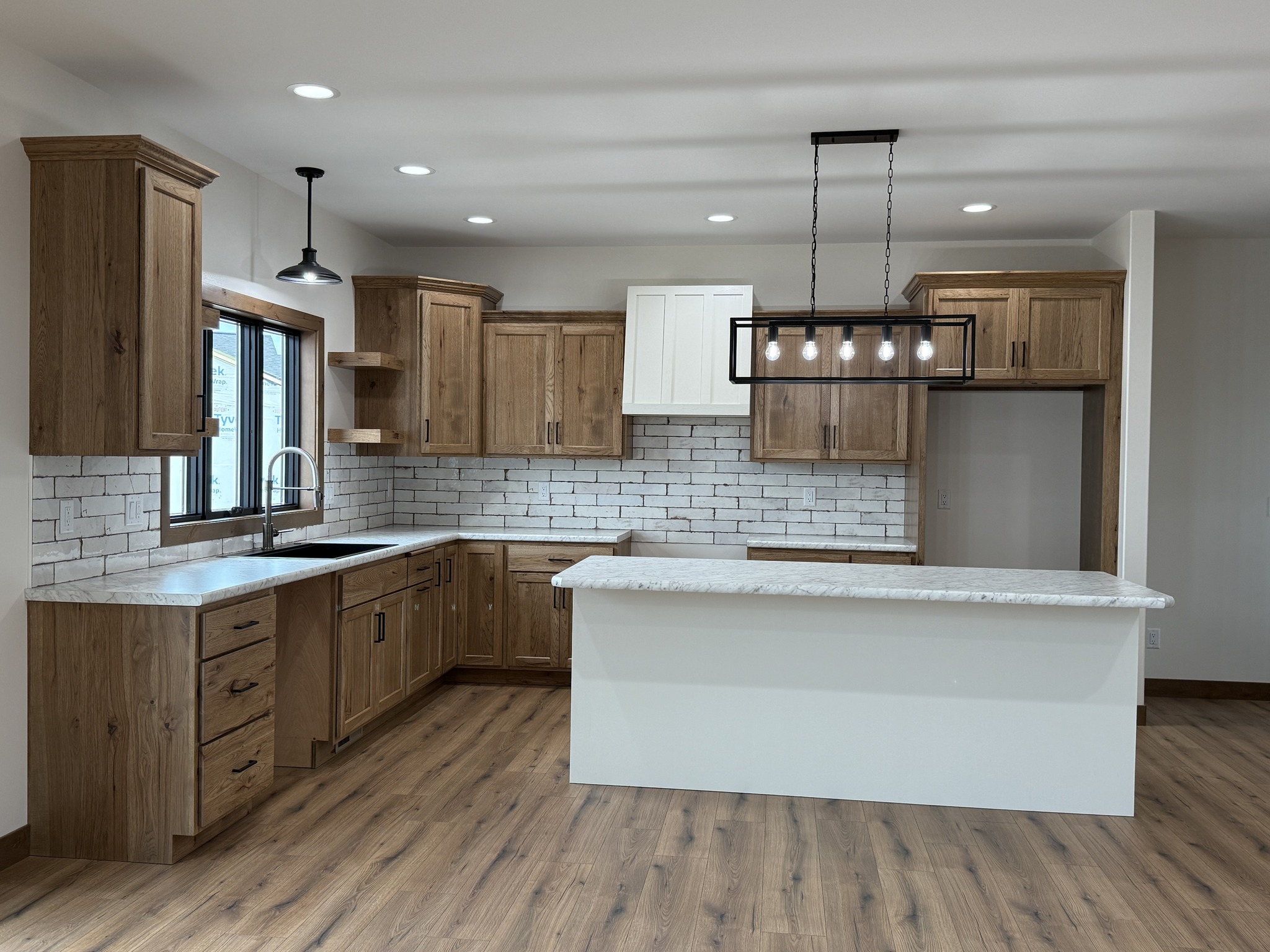 SEO-optimized alt text for a high-ranking modern luxury modular home kitchen featuring professional-grade oak cabinetry and a large white waterfall-style kitchen island with marble countertops; the interior design showcases a trendy white subway tile backsplash, a contemporary black rectangular cage chandelier with Edison bulbs, and premium black hardware, all set against durable wide-plank wood flooring and large black-framed windows that provide abundant natural light, perfectly highlighting luxury prefab home floor plans, gourmet modular kitchen layouts, and high-end residential mobile living finishes for maximum search engine visibility and ranking.