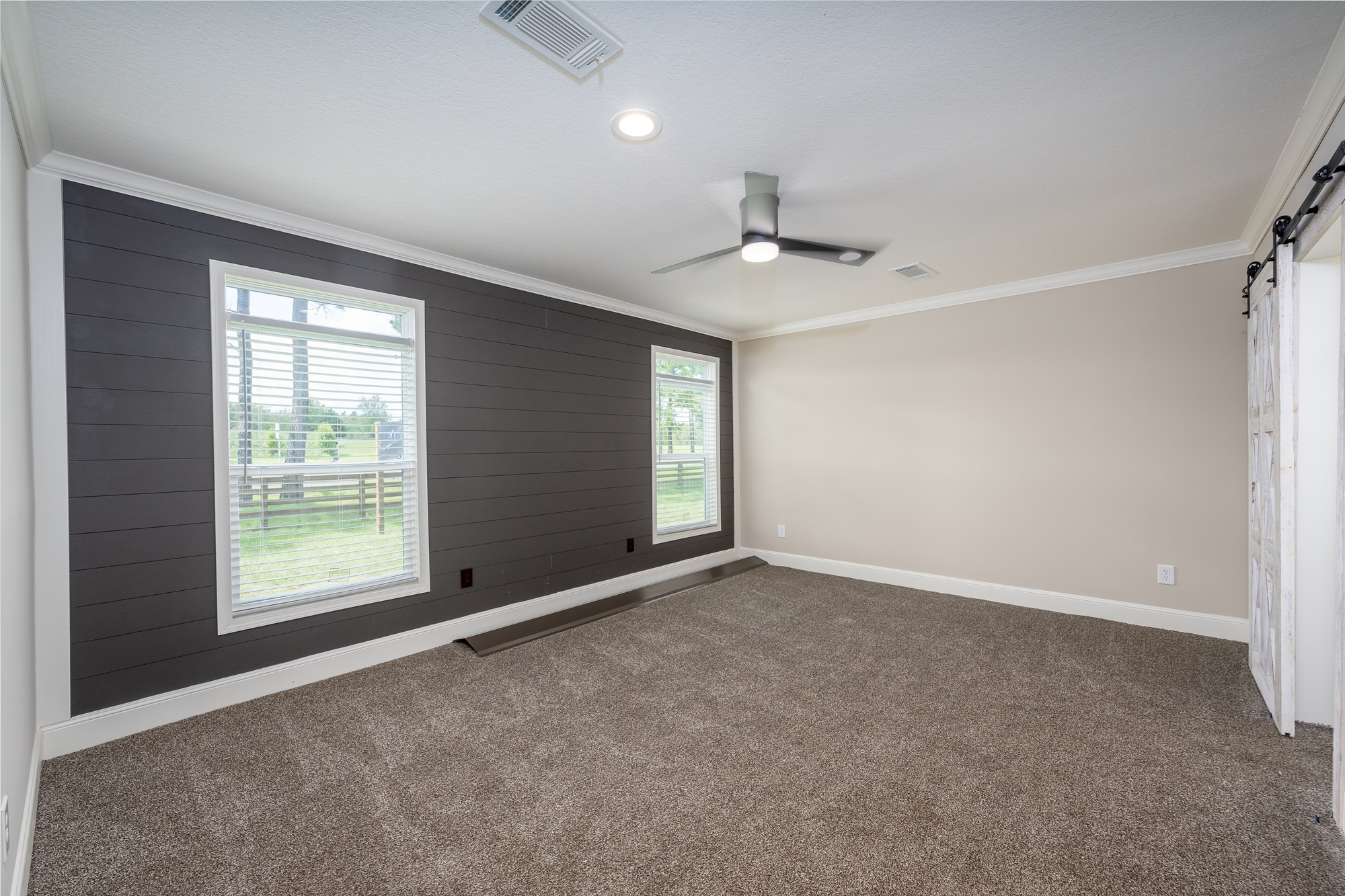 Spacious empty secondary bedroom in The Garnet 1,873 sq ft 3-bedroom customizable modular home featuring modern farmhouse style with dramatic dark gray shiplap accent wall framed in crisp white trim and large double windows with white blinds offering scenic outdoor views of green fields and trees, neutral beige walls and thick plush gray carpet flooring, vaulted textured ceiling with recessed lighting ceiling fan combo for comfort and airflow, rustic white barn door sliding closet entry with black hardware, open bright layout with ample natural light creating a versatile blank canvas ready for personalization as guest room kids bedroom home office or nursery in this dream family home giveaway winner.