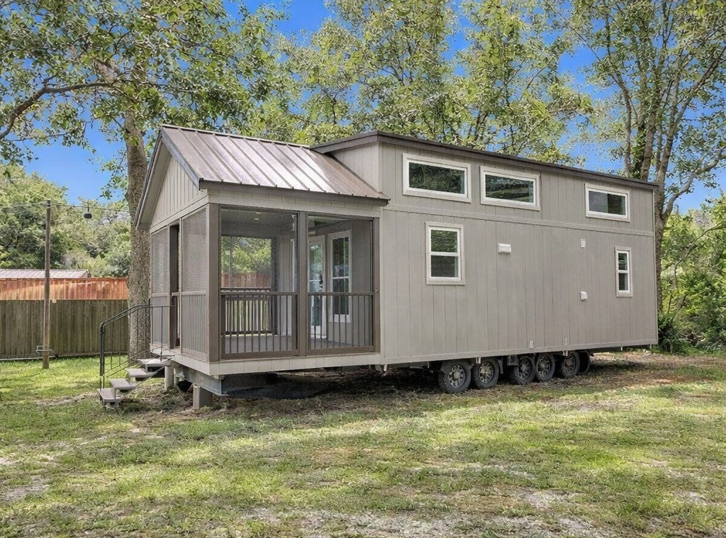 SEO-optimized alt text for a high-ranking luxury custom park model tiny home exterior featuring a professional architectural design with a gabled metal roof and a spacious screened-in front porch; the view showcases a sophisticated taupe prefab residence with vertical board-and-batten siding, a multi-axle chassis for mobility, and contemporary white-framed clerestory windows, perfectly highlighting premium small-space living floor plans, upscale tiny house curb appeal, and luxury residential mobile home finishes for maximum search engine visibility and ranking.