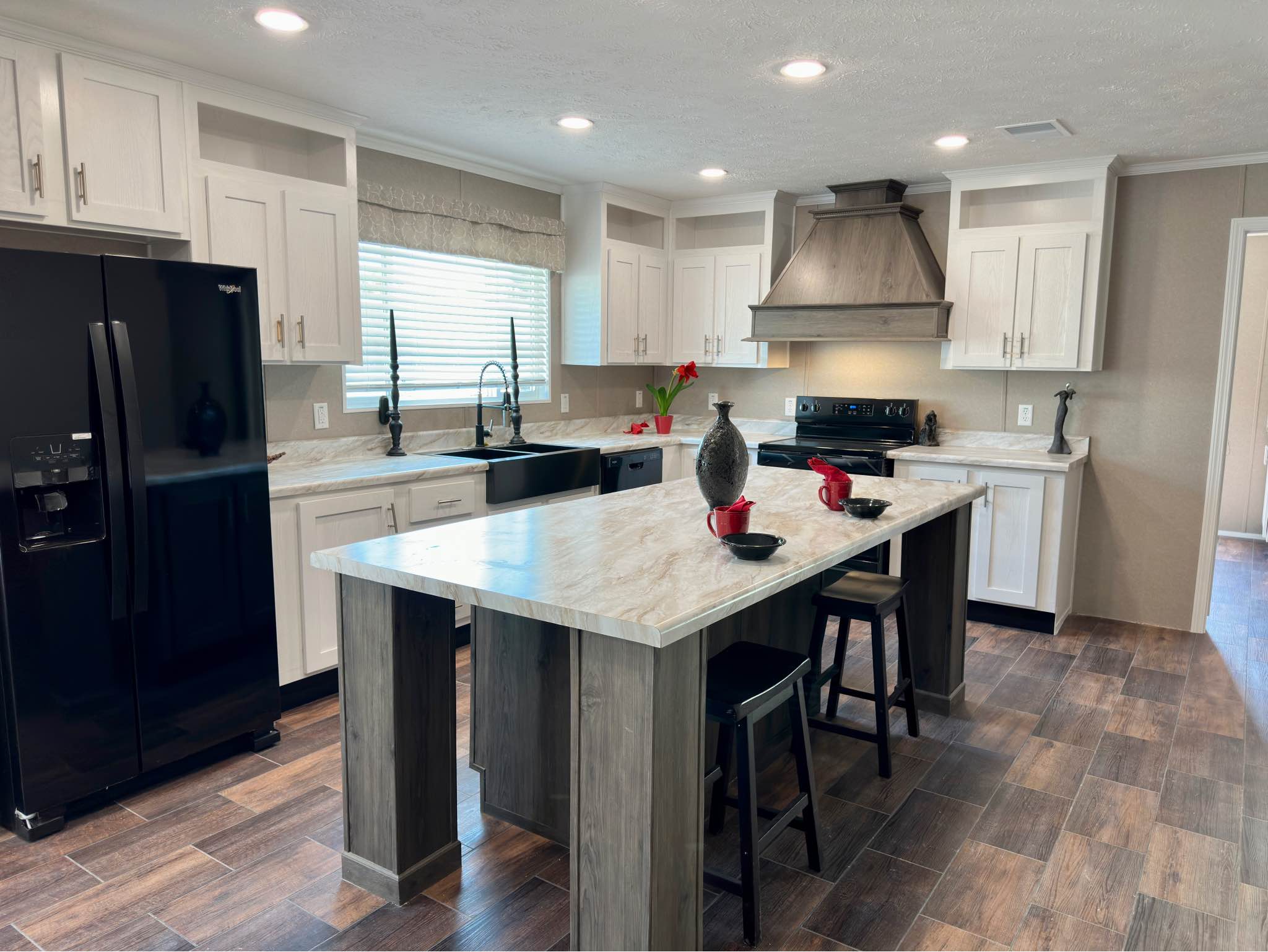 Modern farmhouse kitchen interior featuring a large marble-patterned island with rustic wood legs and black barstools, set against white shaker-style cabinets with matte black hardware, a black farmhouse sink, and high-end black appliances, all illuminated by recessed lighting and complemented by a large rustic wood vent hood and luxury wood-look plank flooring.