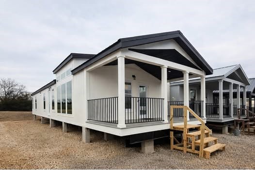 Luxury modern farmhouse tiny home exterior featuring crisp white board and batten siding, dramatic black gable roof accents and trim, spacious wraparound porch with sturdy white columns, black metal railings, vaulted ceiling overhang, expansive floor-to-ceiling panoramic windows flooding interior with natural light, elevated foundation on concrete piers with gravel base, wooden staircase entry, contemporary modular prefab cottage design perfect for rural living, off-grid retreat, vacation home, or family manufactured residence with timeless countryside curb appeal and premium finishes