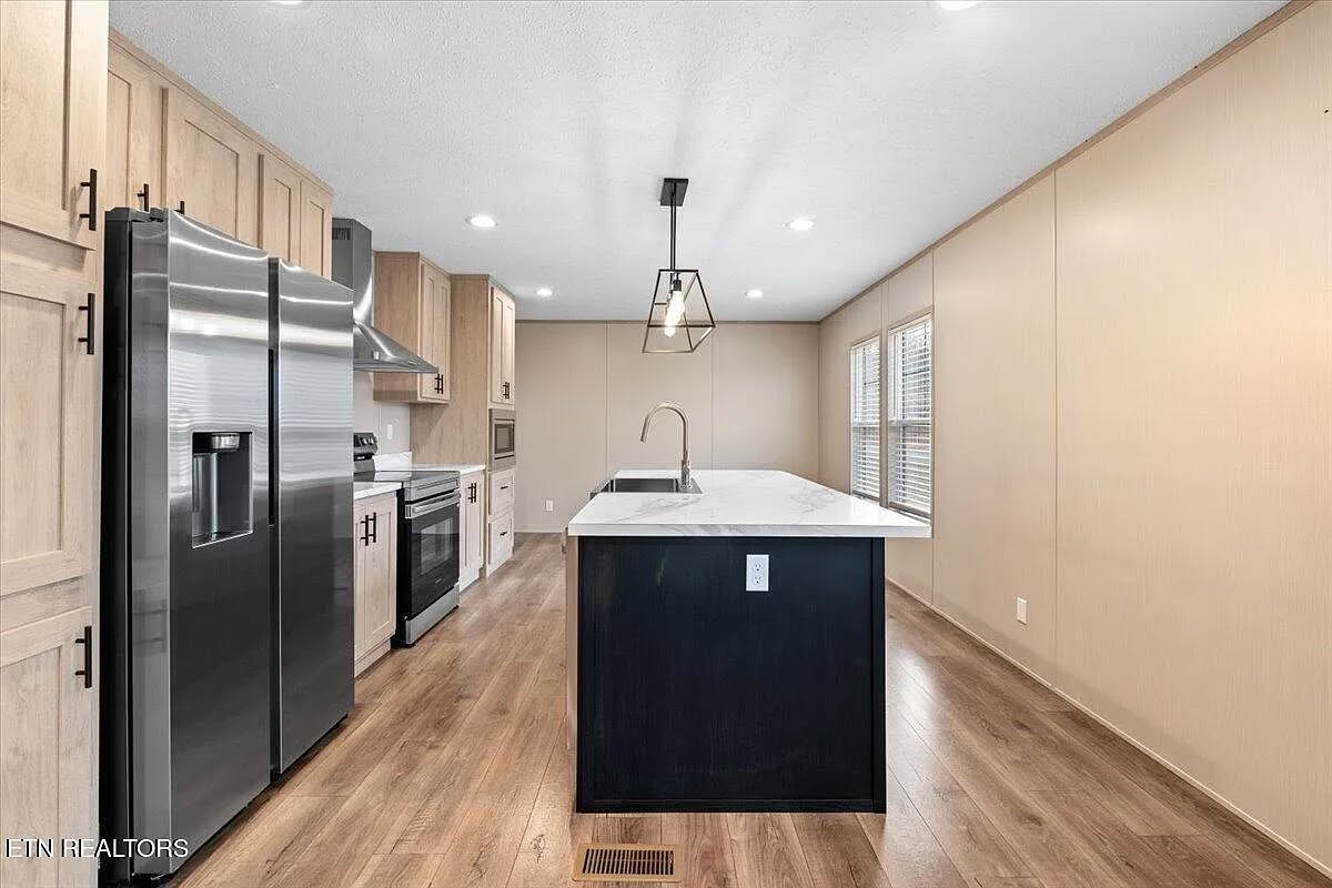 Modern spacious open-concept kitchen interior with large stainless steel double-door refrigerator, light oak shaker cabinets, black island with white marble quartz countertop, farmhouse sink, matte black pendant light, built-in stainless appliances, light wood-look luxury vinyl plank flooring, beige accent walls, recessed lighting, and large windows with blinds – perfect for contemporary modular or manufactured home real estate listing.