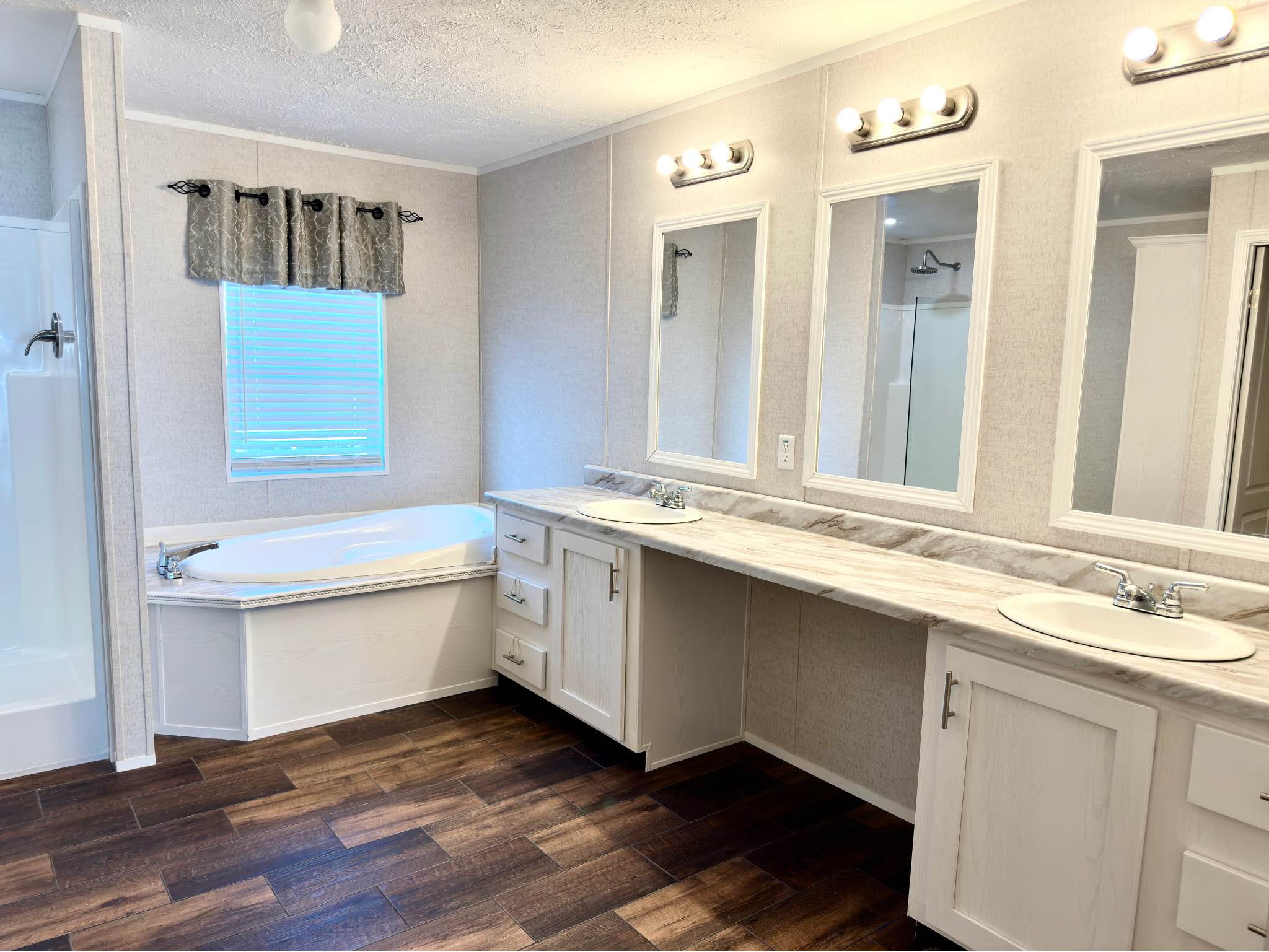 Modern farmhouse master bathroom featuring a luxury double vanity with white cabinetry and marble-style countertops, a large soaking tub nestled under a window with grey valance, and a separate walk-in shower, all tied together by rich dark wood-look plank flooring and bright vanity lighting for a clean, spa-like atmosphere in a contemporary mobile or modular home.
