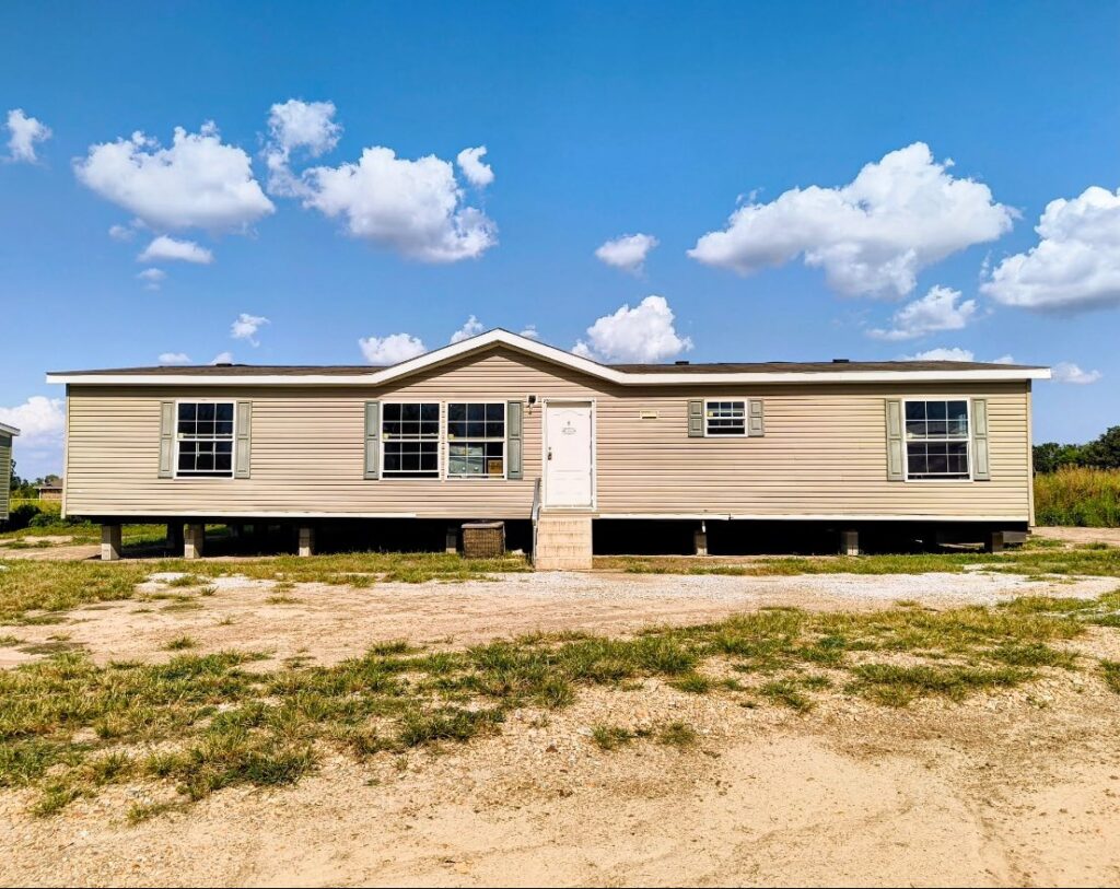 SEO-optimized alt text for a high-end double-wide manufactured home featuring classic beige horizontal vinyl siding, Sage green shutters, and a prominent front gable entry; the long-profile residential modular home is shown on a pier foundation against a clear blue sky with white clouds, highlighting a durable shingle roof and multiple energy-efficient windows, making it ideal for high-ranking searches related to affordable luxury housing, modern mobile home exterior designs, and spacious manufactured home floor plans.