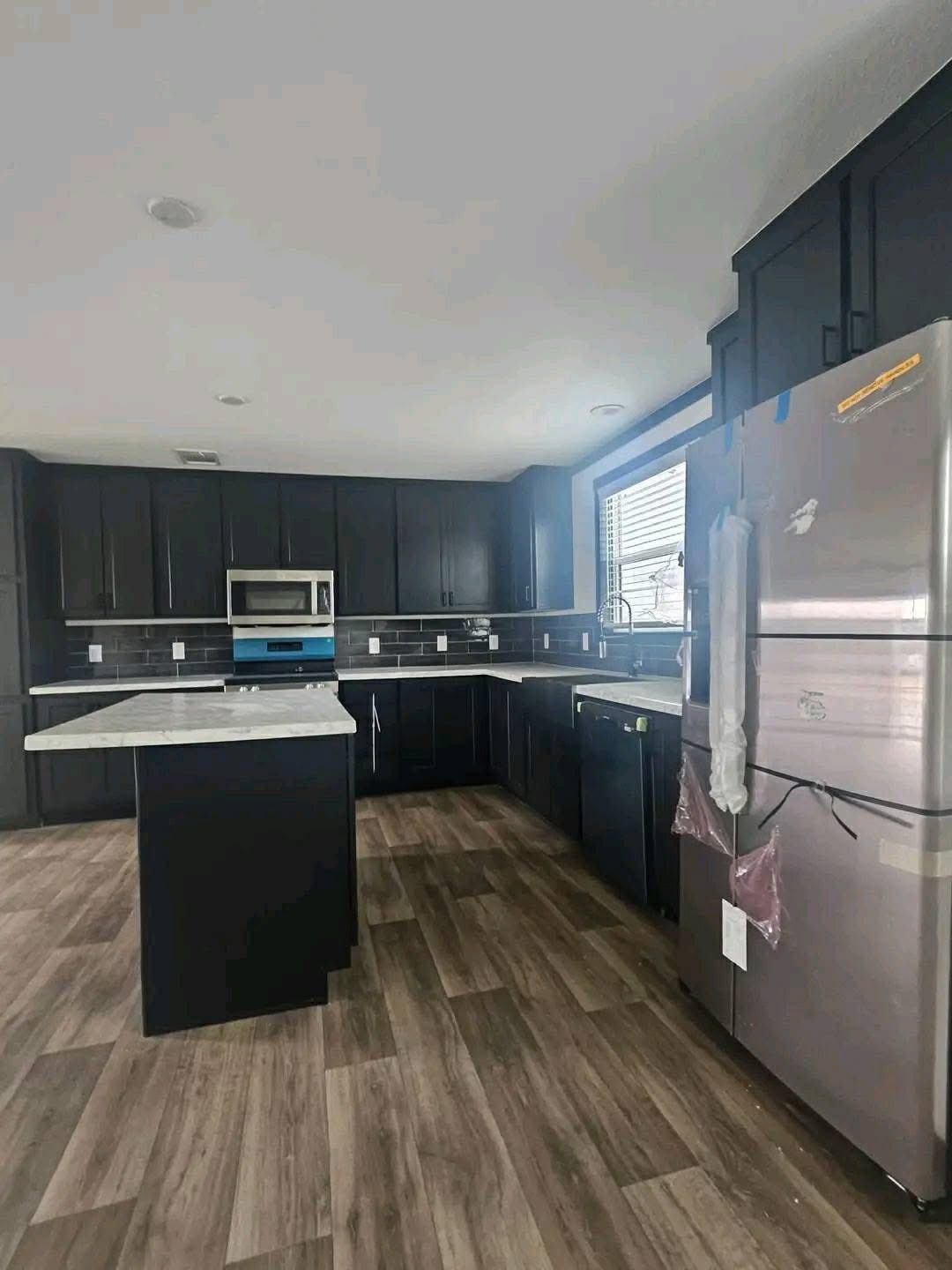 Modern luxury double-wide manufactured home interior featuring a spacious L-shaped kitchen with sleek midnight black cabinetry, a white marble-patterned island with breakfast bar, and high-end stainless steel appliances, all set against a dark subway tile backsplash and premium wide-plank wood-look flooring for a sophisticated 4-bedroom move-in ready turn-key residential design.