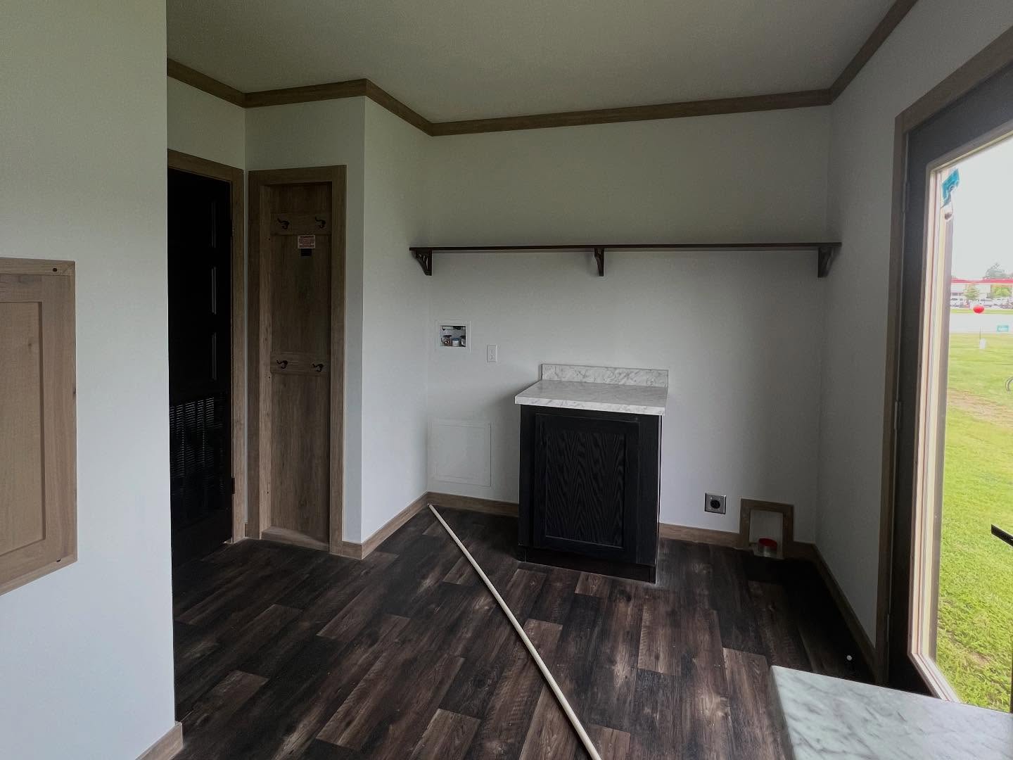 Modern farmhouse laundry room and mudroom entry featuring dark wood-look vinyl flooring, white walls with natural wood trim, and a built-in dark wood cabinet with a marble-style countertop. The functional utility space includes a long wooden hanging shelf, a dedicated recessed coat nook, and a glass-paneled exterior door that overlooks a green lawn, showcasing a practical and stylish transitional area in a contemporary modular or prefab home.