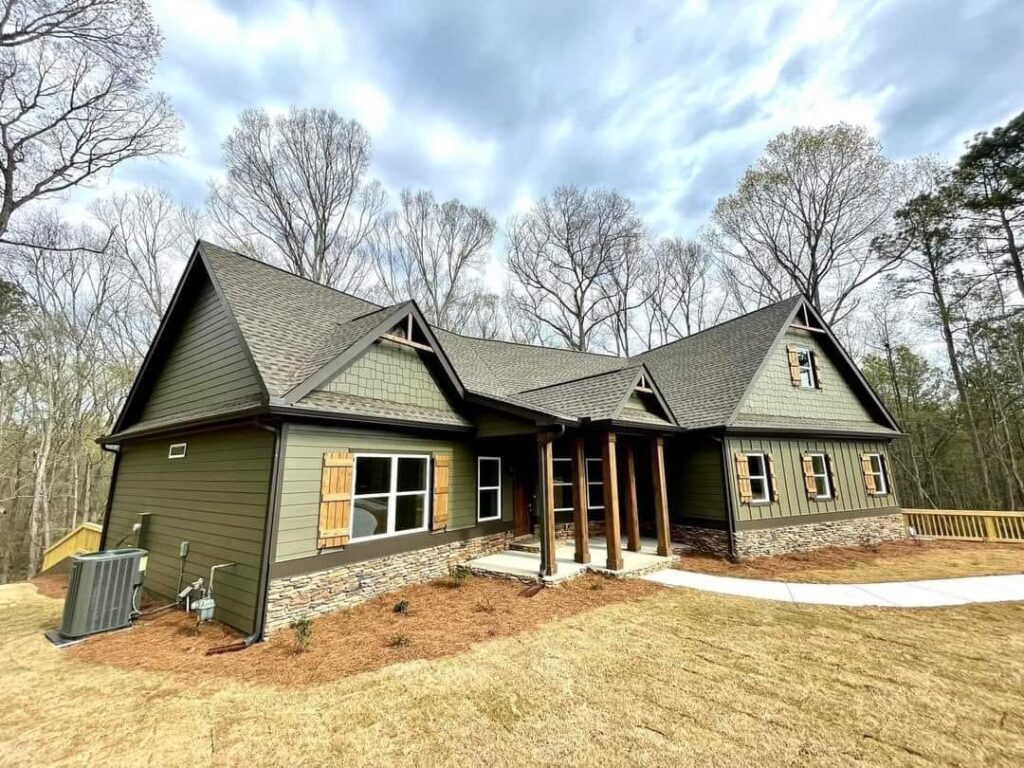 Modern green house with stone accents, wooden shutters, and a sloped roof. Set against tall trees and a cloudy sky, conveying a rustic charm.