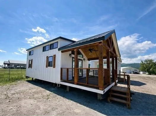SEO-optimized alt text for a high-ranking, modern cabin-style manufactured home featuring a striking white board-and-batten exterior with rustic wood shutters and a spacious covered front porch with natural wood pillars and a gabled roof; the professional architectural view showcases a luxury park model or tiny home design with a dark metal roof and black-trimmed windows, perfectly highlighting premium modular home curb appeal, contemporary portable living solutions, and stylish residential small-space floor plans for maximum search engine visibility and ranking.