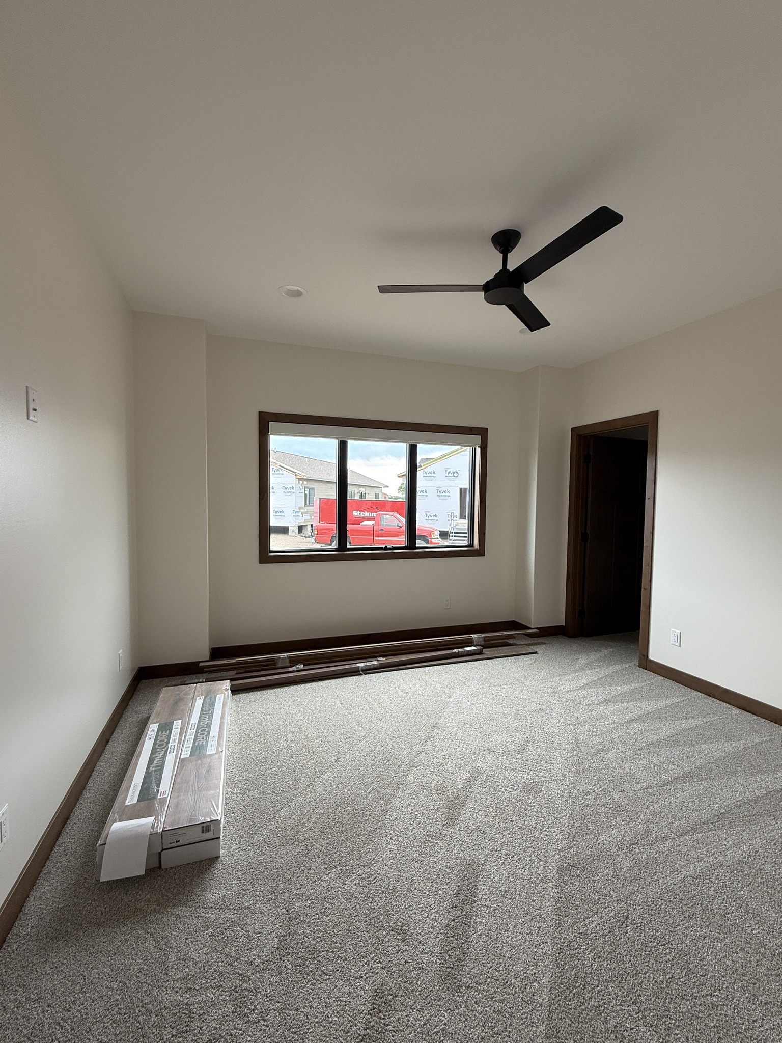 SEO-optimized alt text for a high-ranking modern luxury modular home bedroom featuring a professional interior design with a neutral cream palette and plush beige carpeting; the spacious master suite showcases a contemporary black three-blade ceiling fan, a large black-framed window offering a view of the residential development, and elegant wood-trimmed doorways, perfectly highlighting premium prefab home floor plans, minimalist craftsman-style architectural details, and high-end residential mobile living solutions for maximum search engine visibility and ranking.