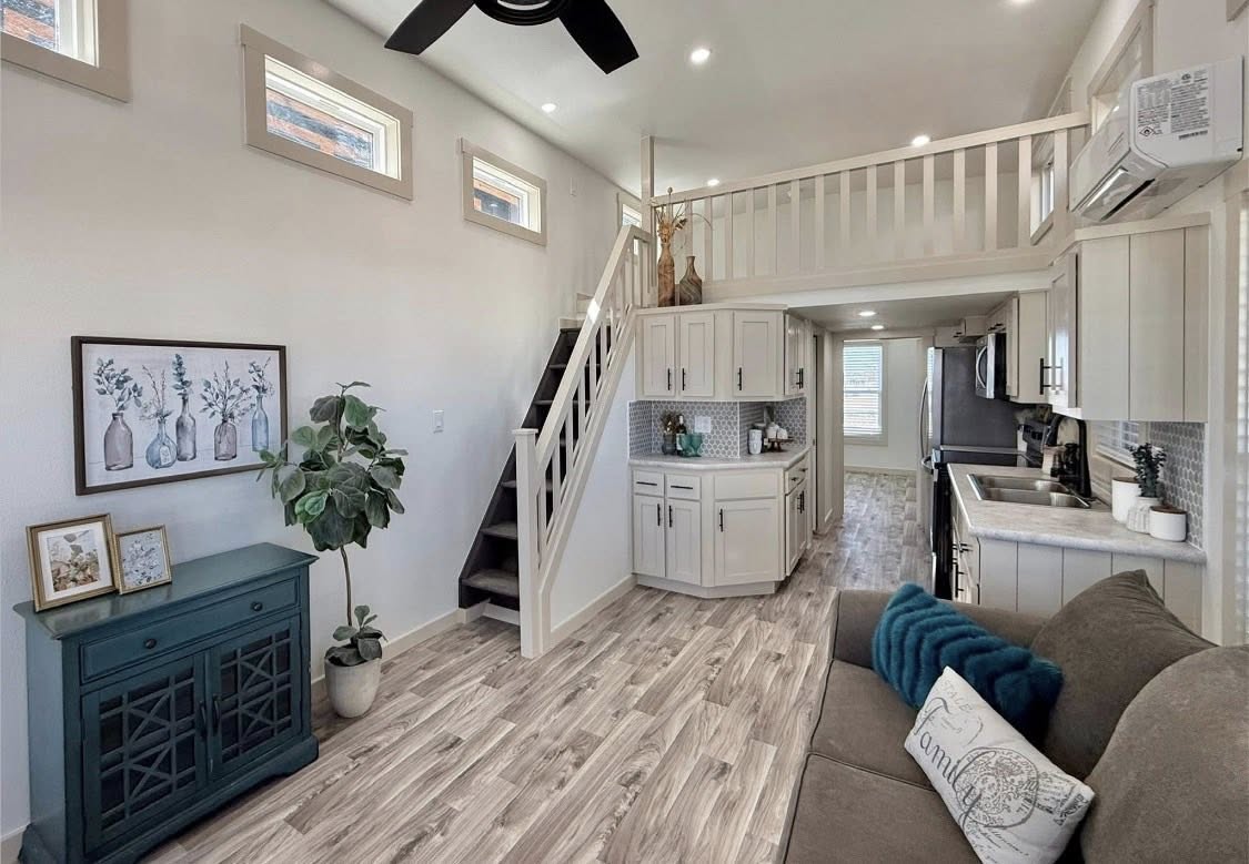 Modern tiny home interior with a cozy loft, wood flooring, stylish kitchen, teal cabinet, and stairs. Bright and welcoming atmosphere.